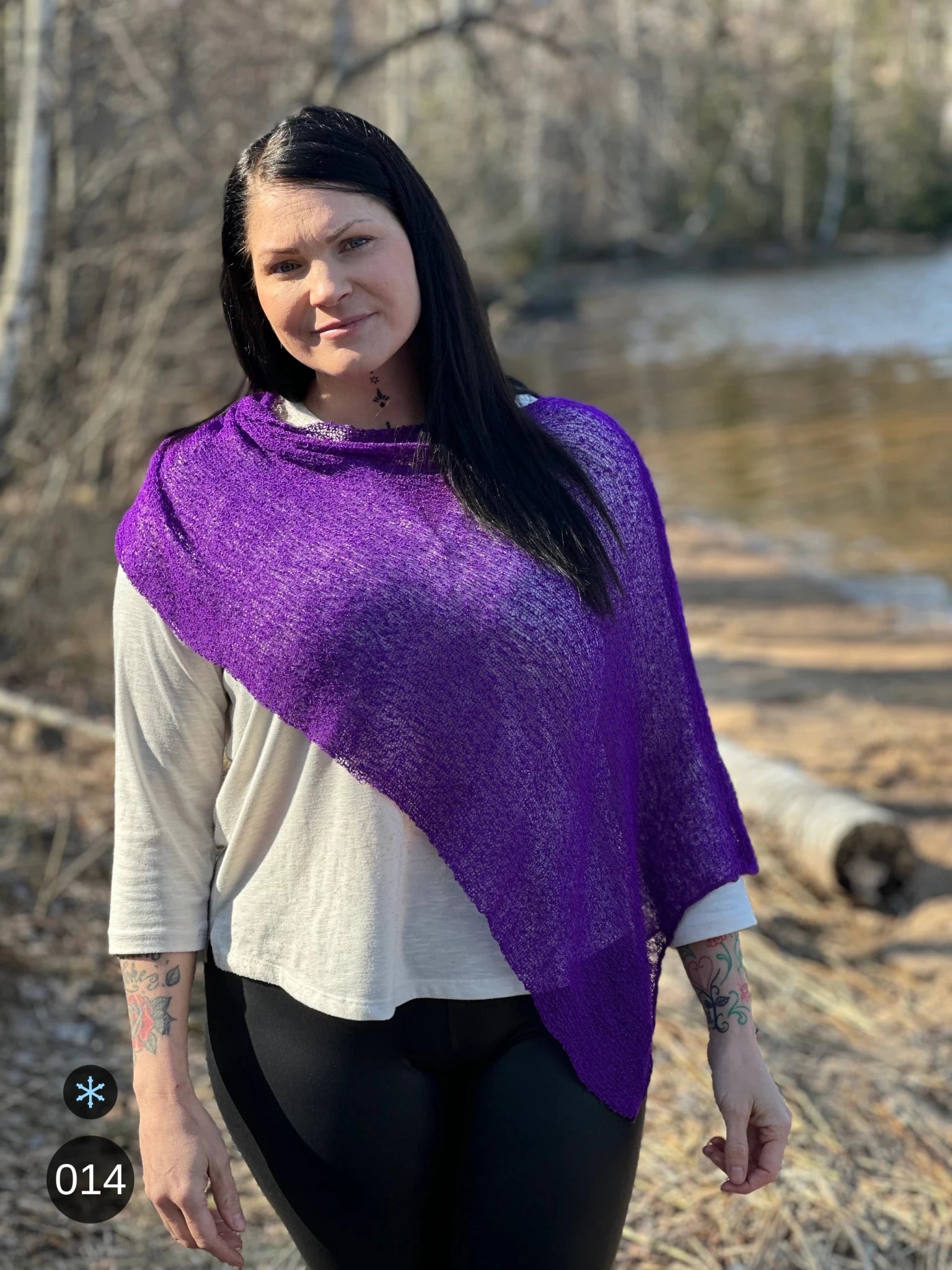Kvinna bär Ella Cape i färg 014 Mörklila, en kall färg i djup violett lila, finstickad cape som draperas diagonalt över ena skuldran, fotad vid en å i vårljus. Ulla Jacobsson Kläder.