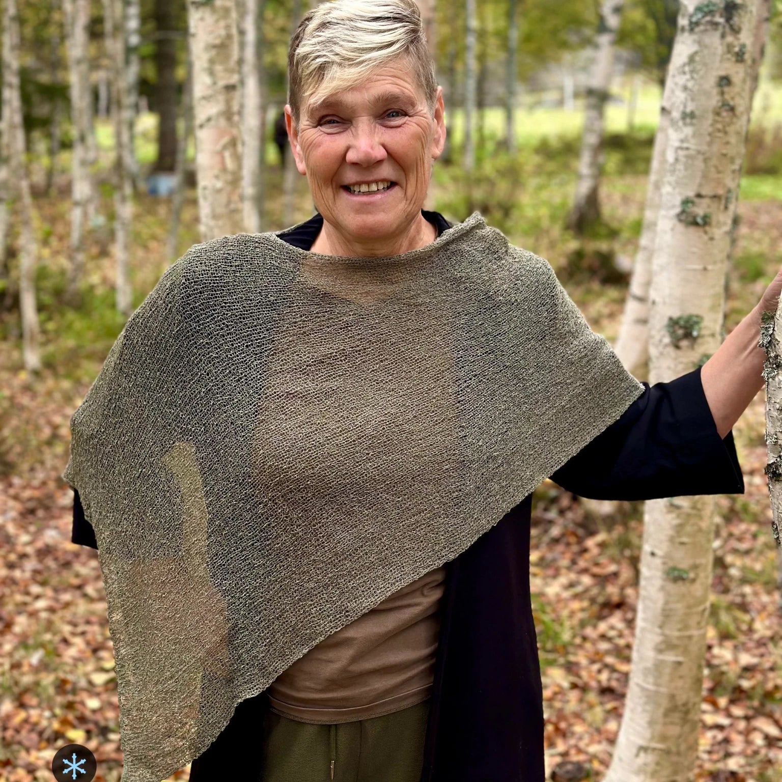 Kvinna bär Ella Cape i färg 016 Lavgrön, en kall färg i dämpad gråbrun grönton som lav på sten, finstickad cape som draperas mjukt över axlarna, fotad vid en björkstam i höstsol. Ulla Jacobsson Kläder.