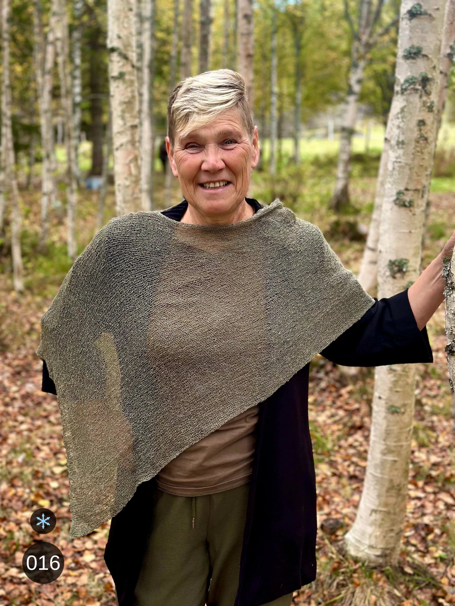 Kvinna bär Ella Cape i färg 016 Lavgrön, en kall färg i dämpad gråbrun grönton som lav på sten, finstickad cape som draperas mjukt över axlarna, fotad vid en björkstam i höstsol. Ulla Jacobsson Kläder.