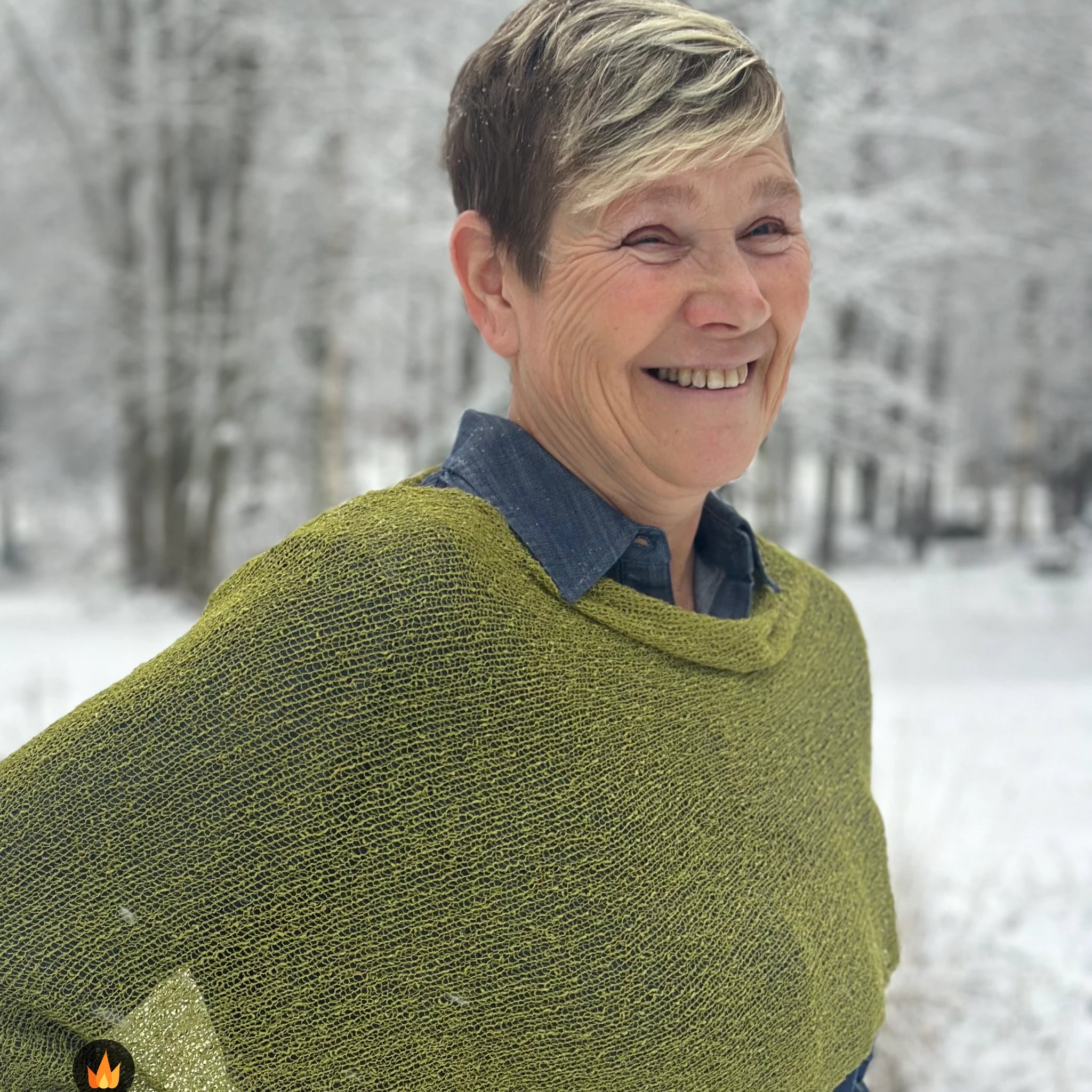 Kvinna bär Ella Cape i färg 017 Olivgrön, en varm färg i mättad olivgrön, finstickad cape som draperas mjukt över axlarna, fotad i ett frostigt vinterlandskap. Ulla Jacobsson Kläder.