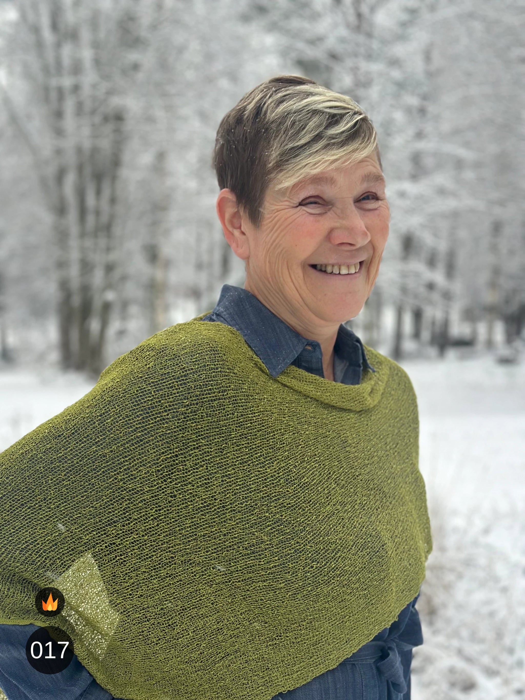 Kvinna bär Ella Cape i färg 017 Olivgrön, en varm färg i mättad olivgrön, finstickad cape som draperas mjukt över axlarna, fotad i ett frostigt vinterlandskap. Ulla Jacobsson Kläder.
