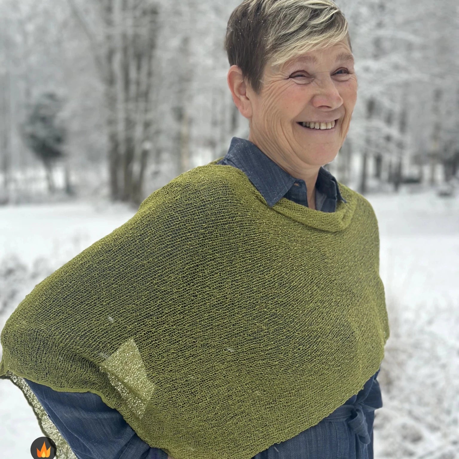Kvinna bär Ella Cape i färg 017 Olivgrön, en varm färg, finstickad cape som draperas mjukt över axlarna, fotad i ett frostigt vinterlandskap. Ulla Jacobsson Kläder.