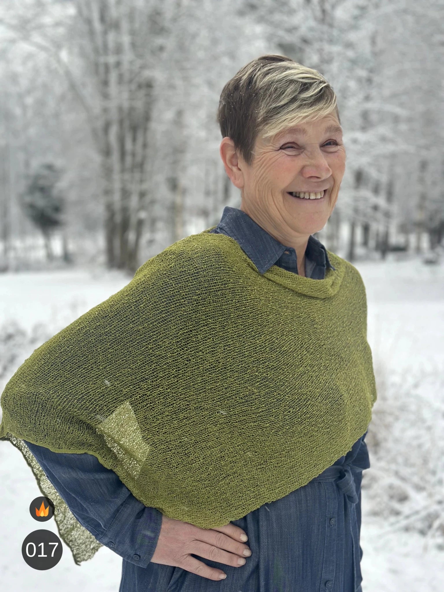 Kvinna bär Ella Cape i färg 017 Olivgrön, en varm färg, finstickad cape som draperas mjukt över axlarna, fotad i ett frostigt vinterlandskap. Ulla Jacobsson Kläder.