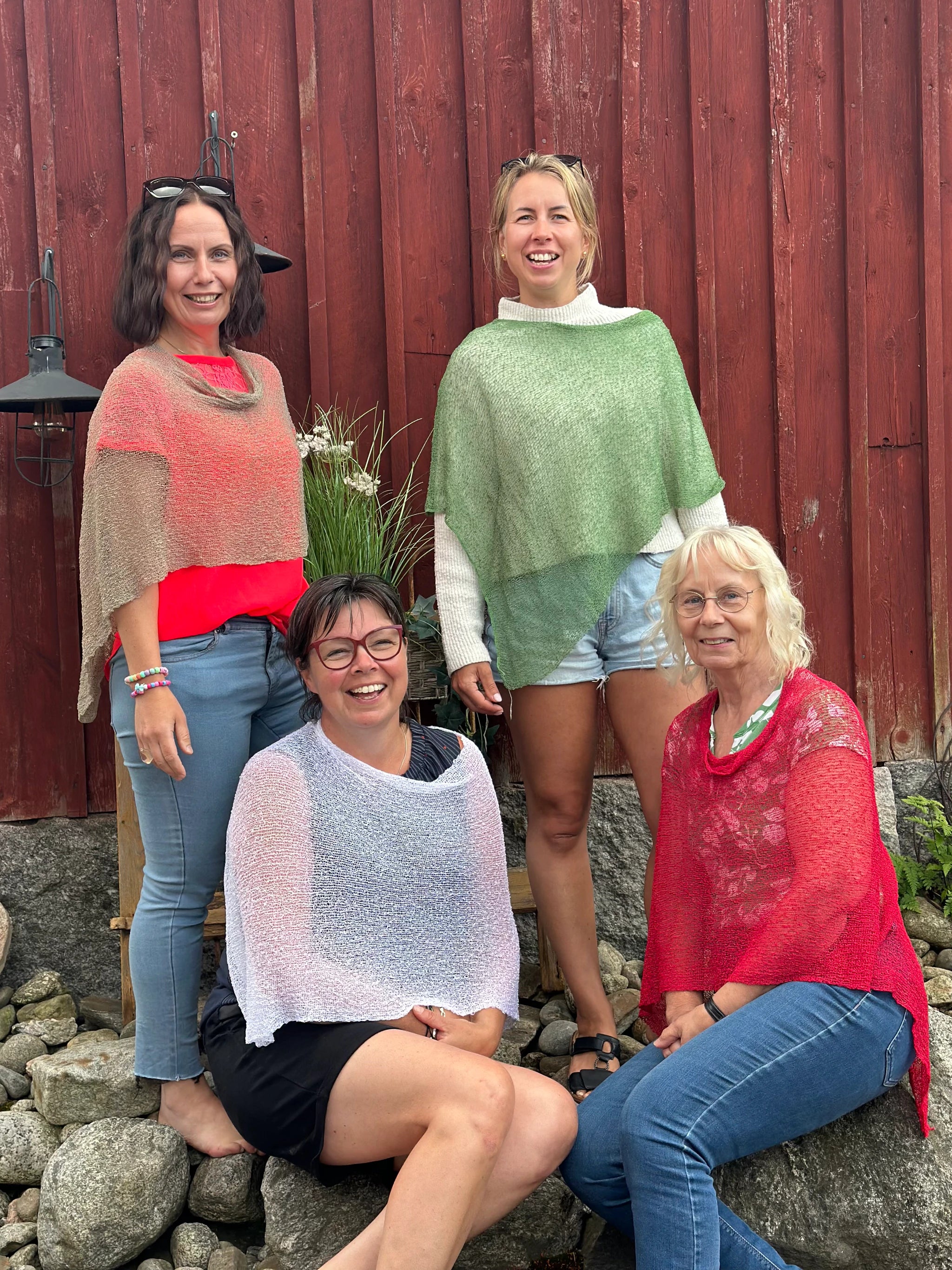 Fyra kvinnor bär Ella Cape i fyra färger: 123 Ljusbeige, 034 Ljusgrön, 019 Vit och 042 Röd. Finstickade capes som draperas mjukt över axlarna, fotade mot en faluröd timmervägg en sommardag. Ulla Jacobsson Kläder.