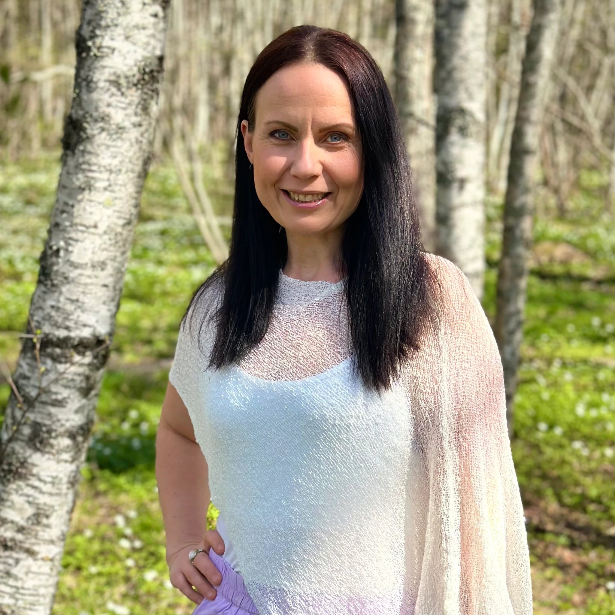 Kvinna bär Ella Cape i färg 019 Vit, en neutral ren vit med mjuk lyster, finstickad cape som draperas mjukt över axlarna, fotad i en björkdunge full av vitsippor en vårdag. Ulla Jacobsson Kläder.