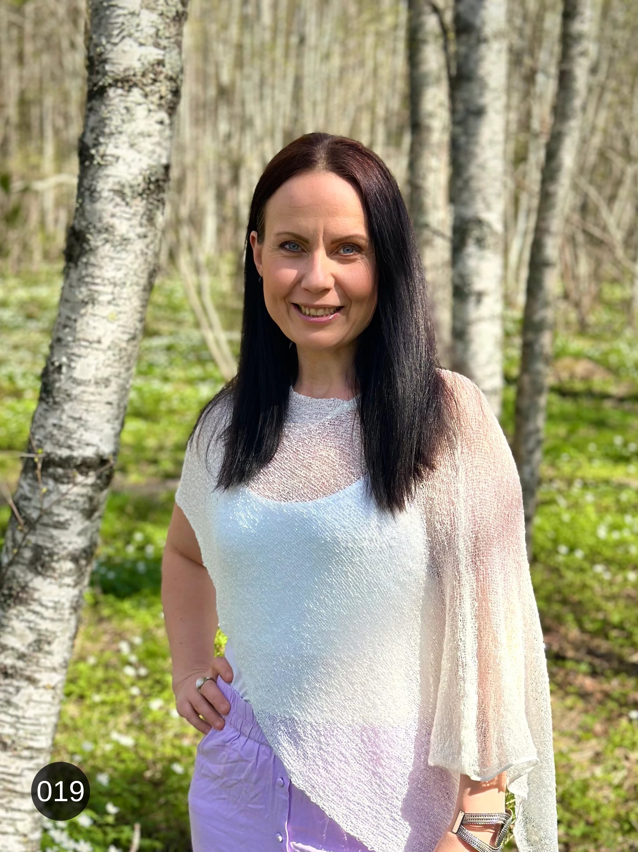Kvinna bär Ella Cape i färg 019 Vit, en neutral ren vit med mjuk lyster, finstickad cape som draperas mjukt över axlarna, fotad i en björkdunge full av vitsippor en vårdag. Ulla Jacobsson Kläder.