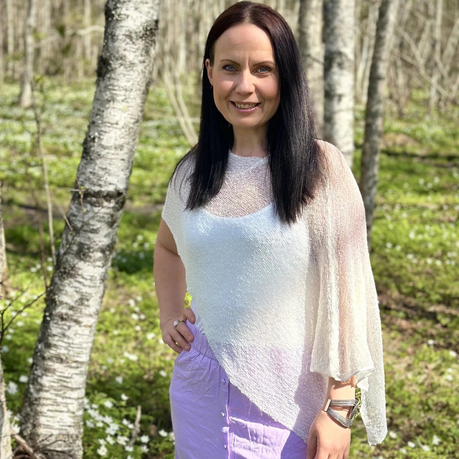 Kvinna bär Ella Cape i färg 019 Vit, finstickad cape som draperas mjukt över axlarna, fotad i en björkdunge med vitsippor. Ulla Jacobsson Kläder.