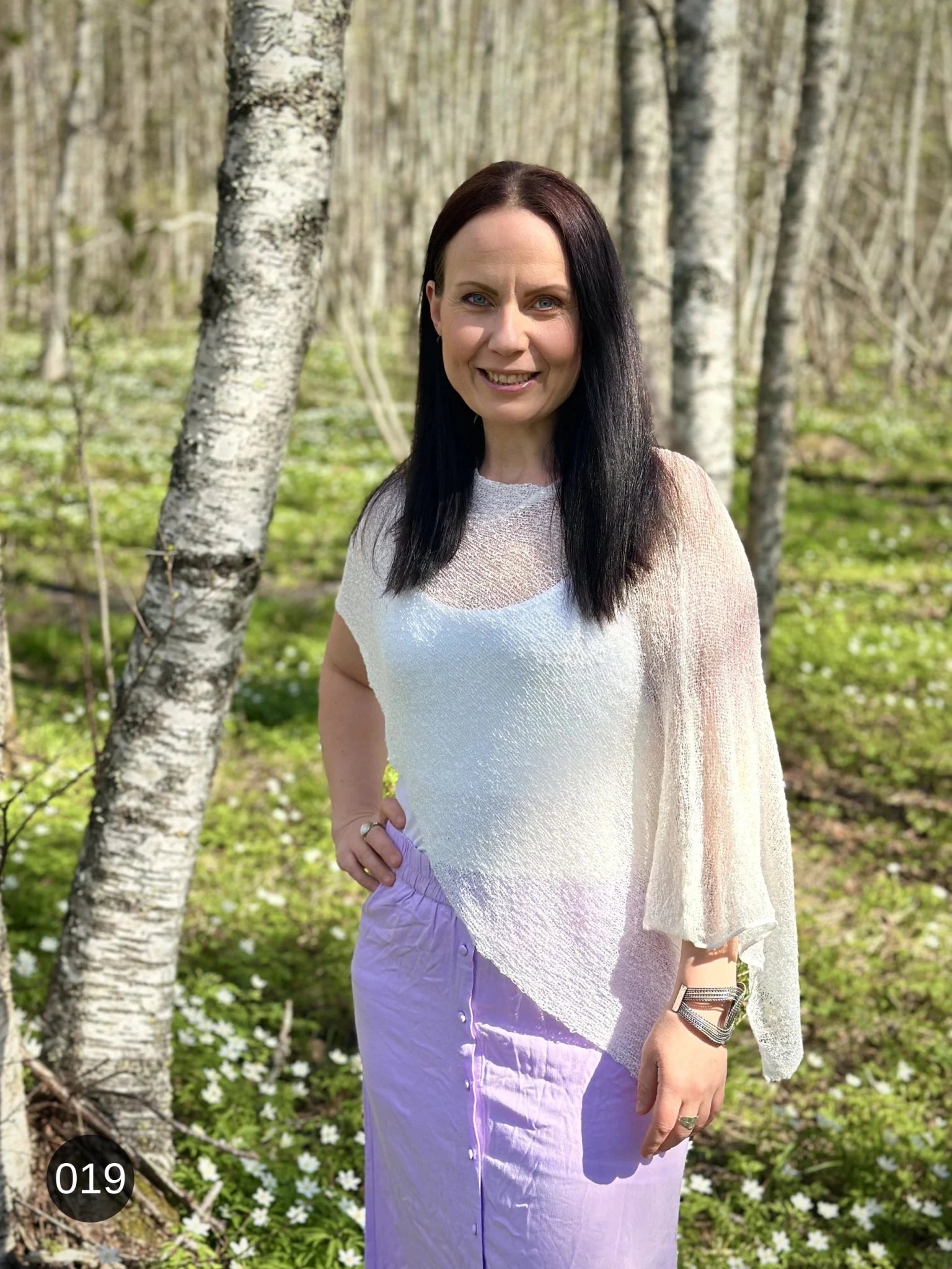 Kvinna bär Ella Cape i färg 019 Vit, finstickad cape som draperas mjukt över axlarna, fotad i en björkdunge med vitsippor. Ulla Jacobsson Kläder.