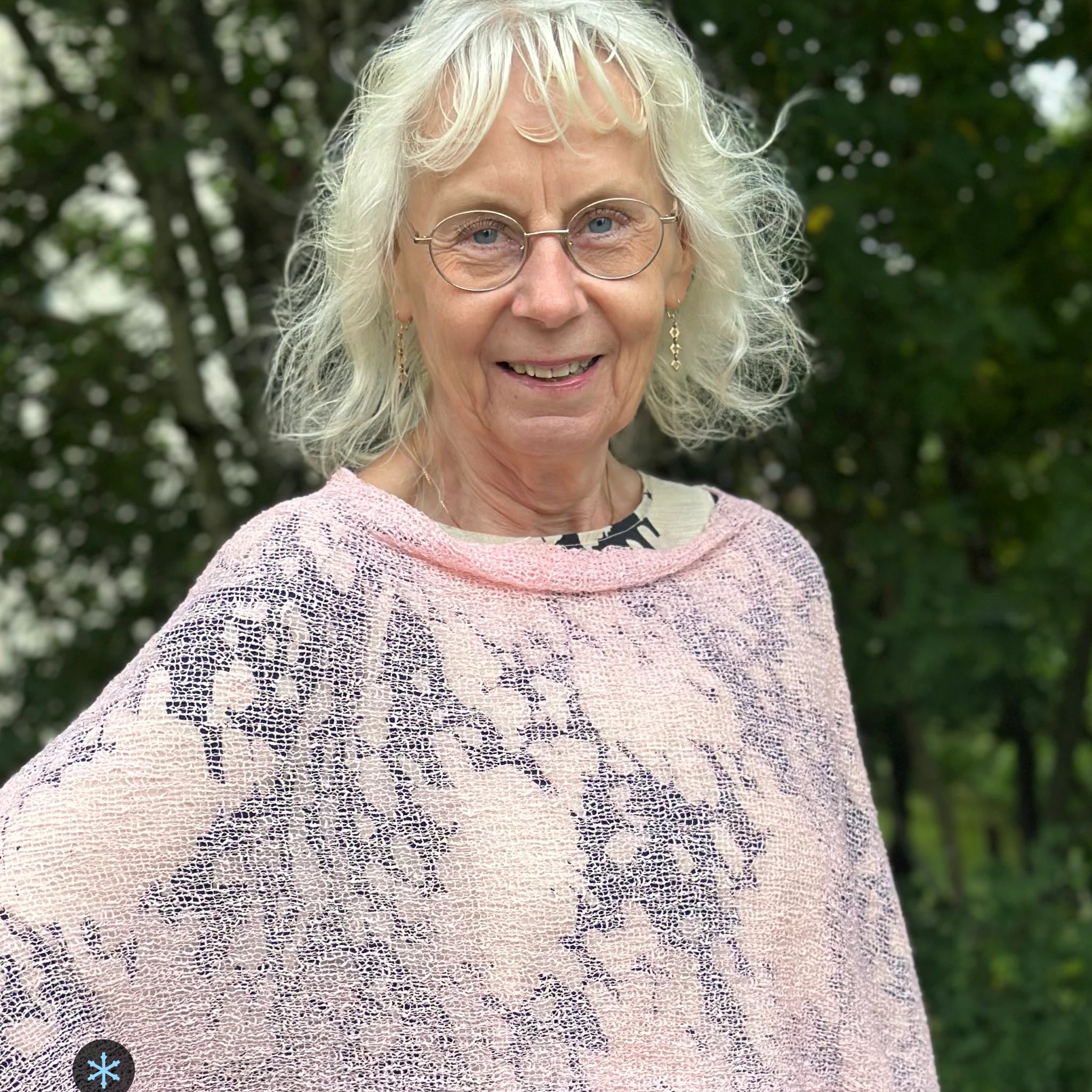Kvinna bär Ella Cape i färg 020 Tuggummirosa, en kall färg i ljus tuggummirosa, finstickad cape som draperas mjukt över axlarna, fotad utomhus i grönska. Ulla Jacobsson Kläder.