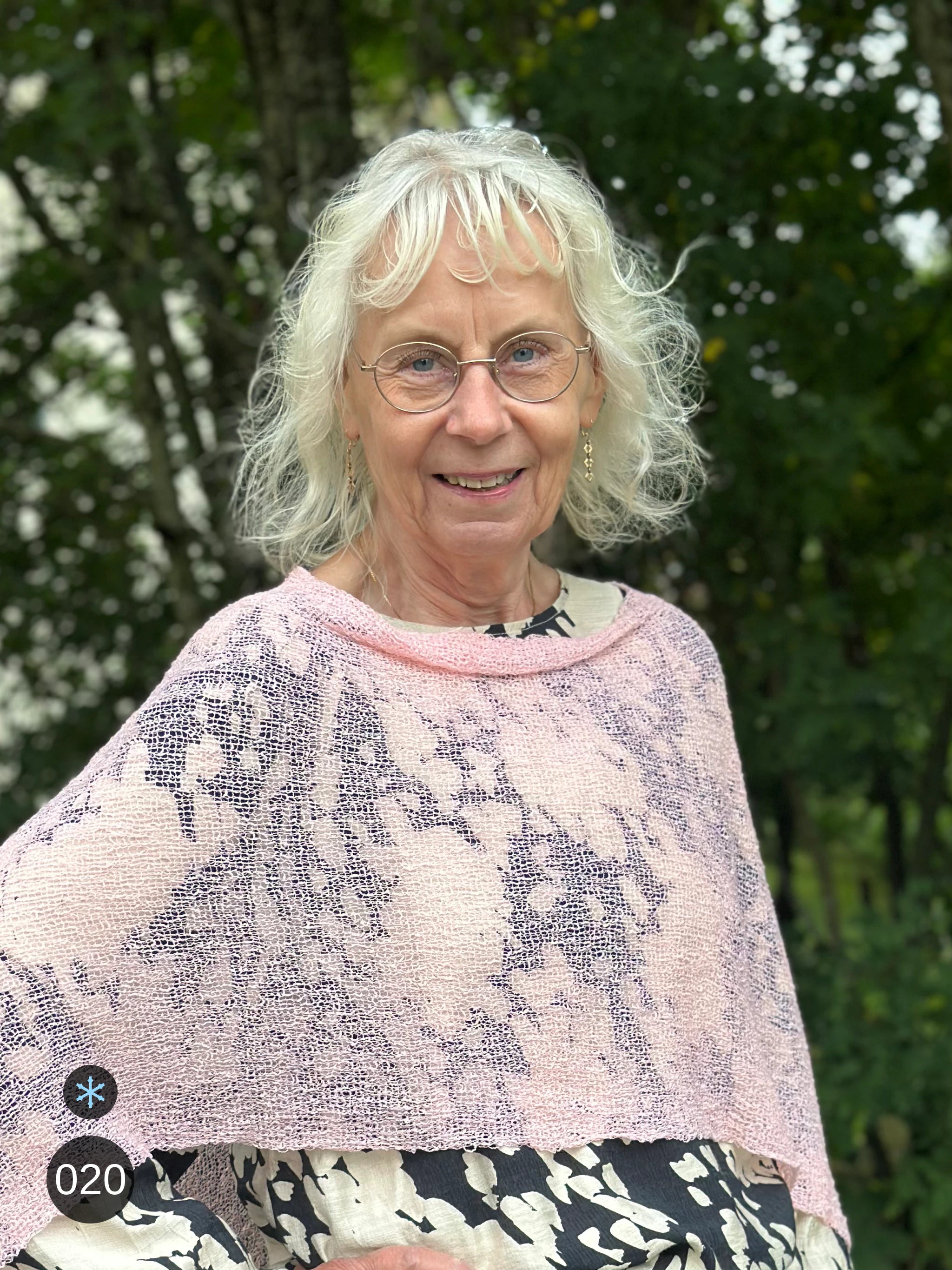 Kvinna bär Ella Cape i färg 020 Tuggummirosa, en kall färg i ljus tuggummirosa, finstickad cape som draperas mjukt över axlarna, fotad utomhus i grönska. Ulla Jacobsson Kläder.