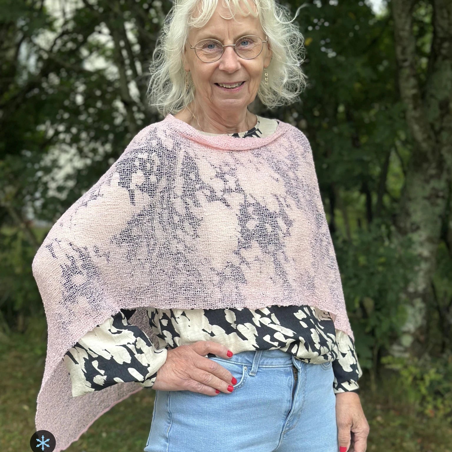 Kvinna bär Ella Cape i färg 020 Tuggummirosa, en kall färg, finstickad cape som draperas mjukt över axlarna, fotad utomhus i grönska. Ulla Jacobsson Kläder.