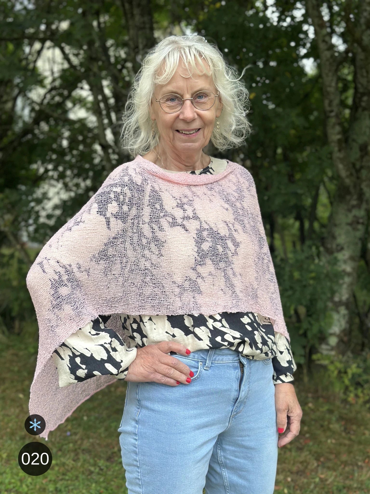 Kvinna bär Ella Cape i färg 020 Tuggummirosa, en kall färg, finstickad cape som draperas mjukt över axlarna, fotad utomhus i grönska. Ulla Jacobsson Kläder.