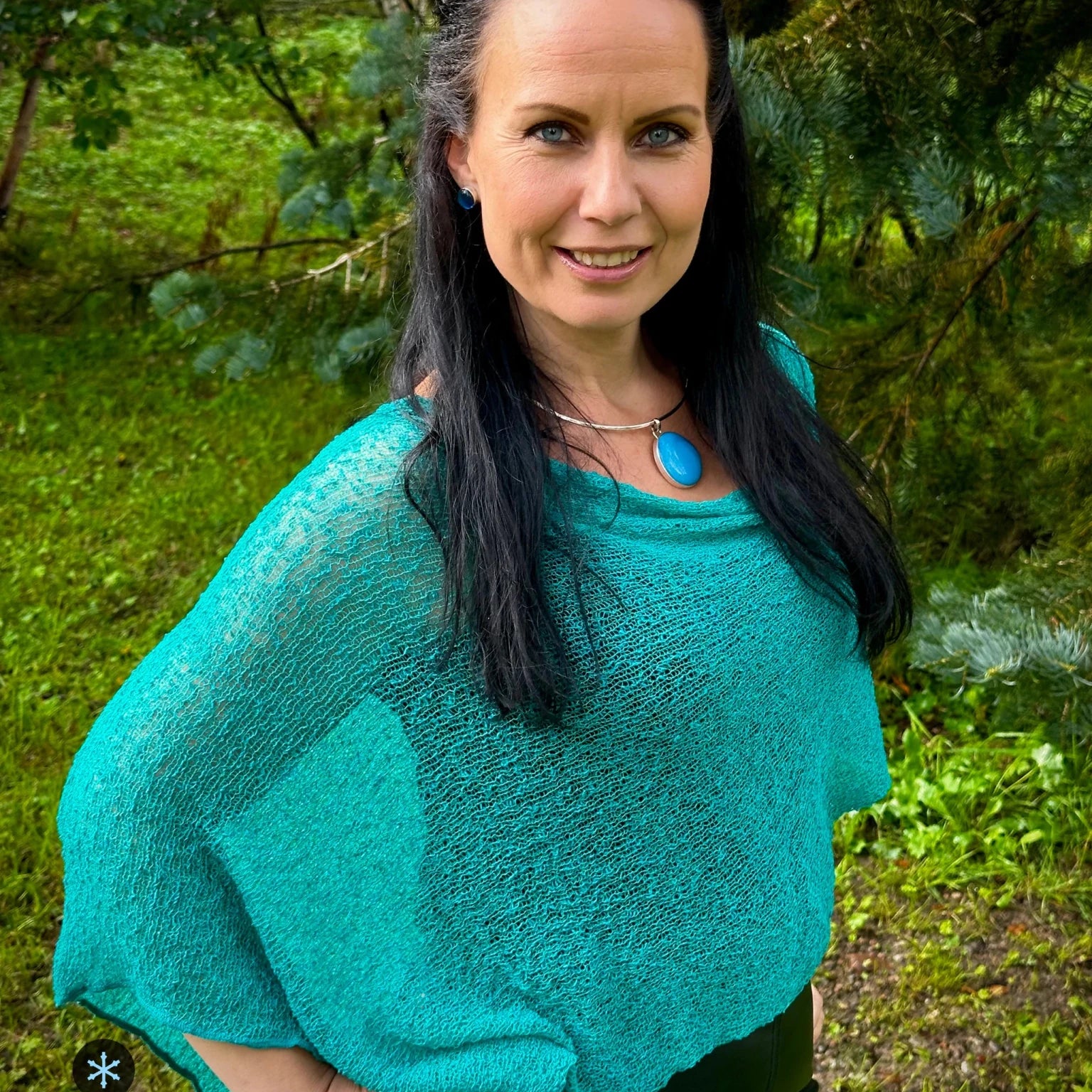 Kvinna bär Ella Cape i färg 021 Grönturkos, en kall färg i klar blågrön turkos, finstickad cape som draperas mjukt över axlarna, fotad i grönskande trädgård. Ulla Jacobsson Kläder.