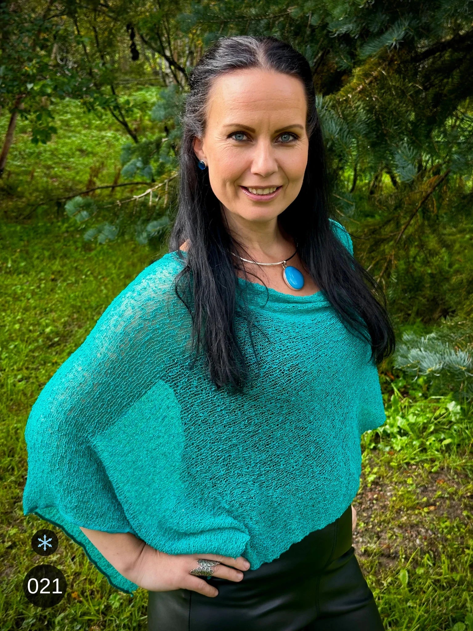 Kvinna bär Ella Cape i färg 021 Grönturkos, en kall färg i klar blågrön turkos, finstickad cape som draperas mjukt över axlarna, fotad i grönskande trädgård. Ulla Jacobsson Kläder.