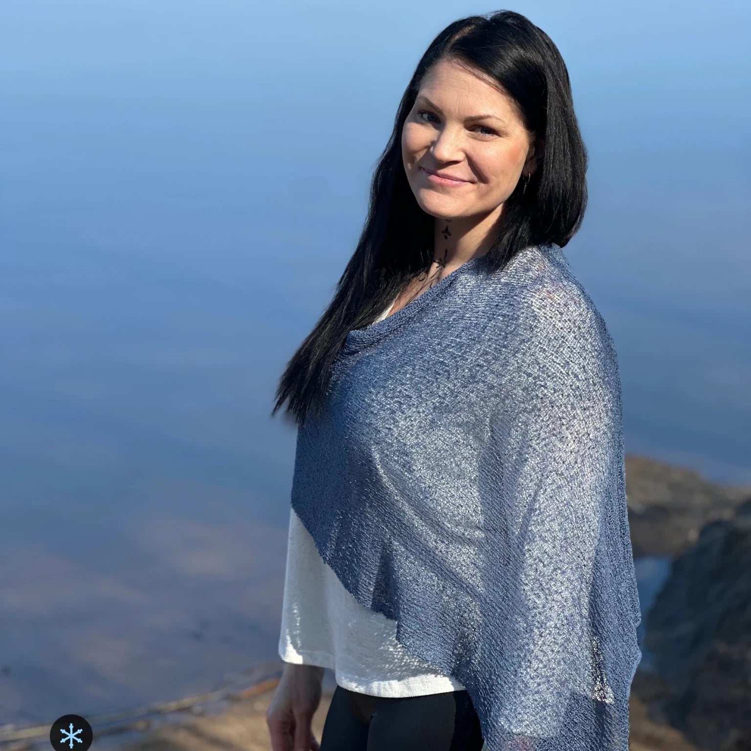 Kvinna bär Ella Cape i färg 025 Jeansblå, en kall färg i dämpad denimblå, finstickad cape som draperas mjukt över axlarna, fotad vid vattnet i kvällsljus. Ulla Jacobsson Kläder.