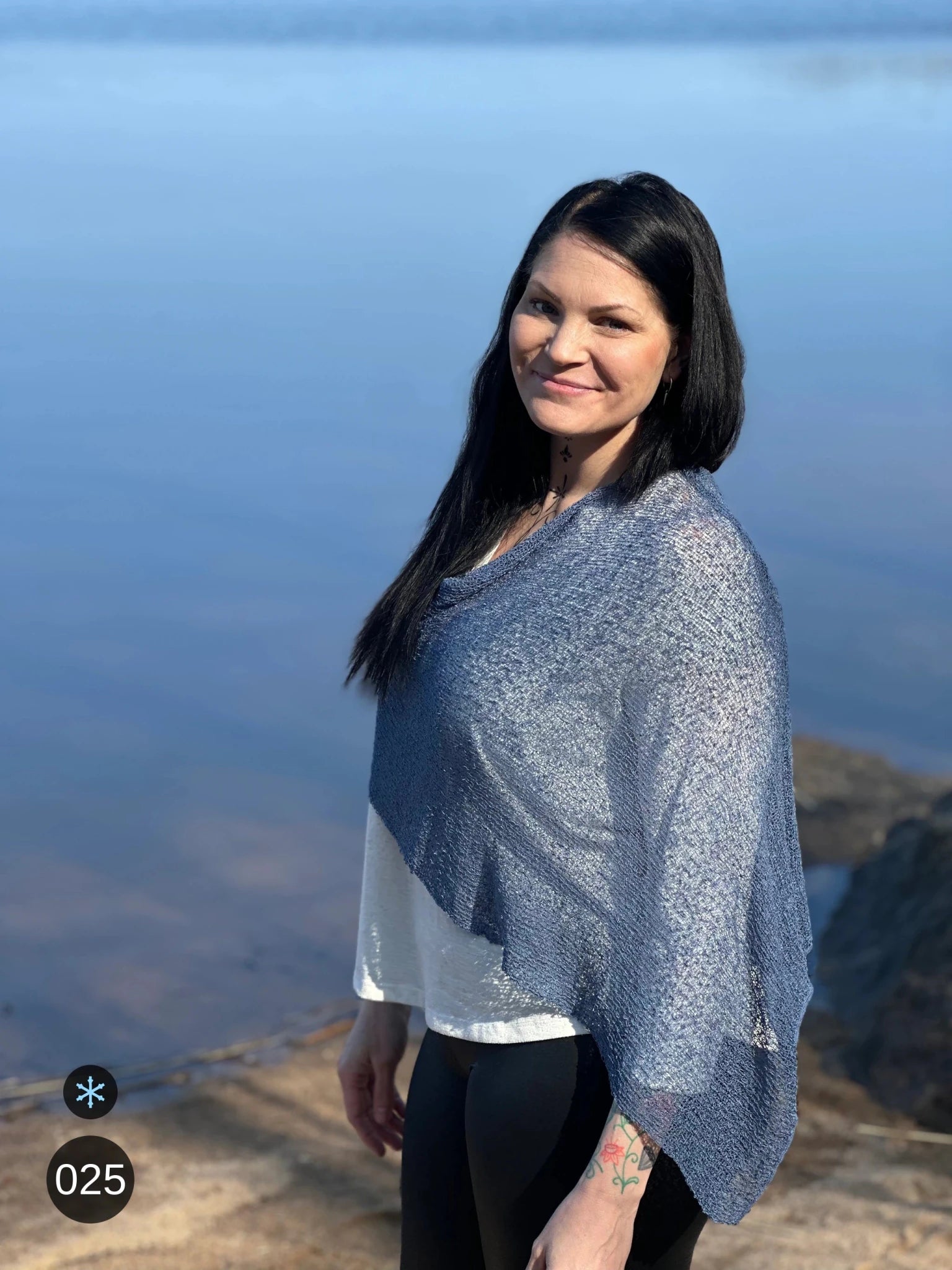 Kvinna bär Ella Cape i färg 025 Jeansblå, en kall färg i dämpad denimblå, finstickad cape som draperas mjukt över axlarna, fotad vid vattnet i kvällsljus. Ulla Jacobsson Kläder.