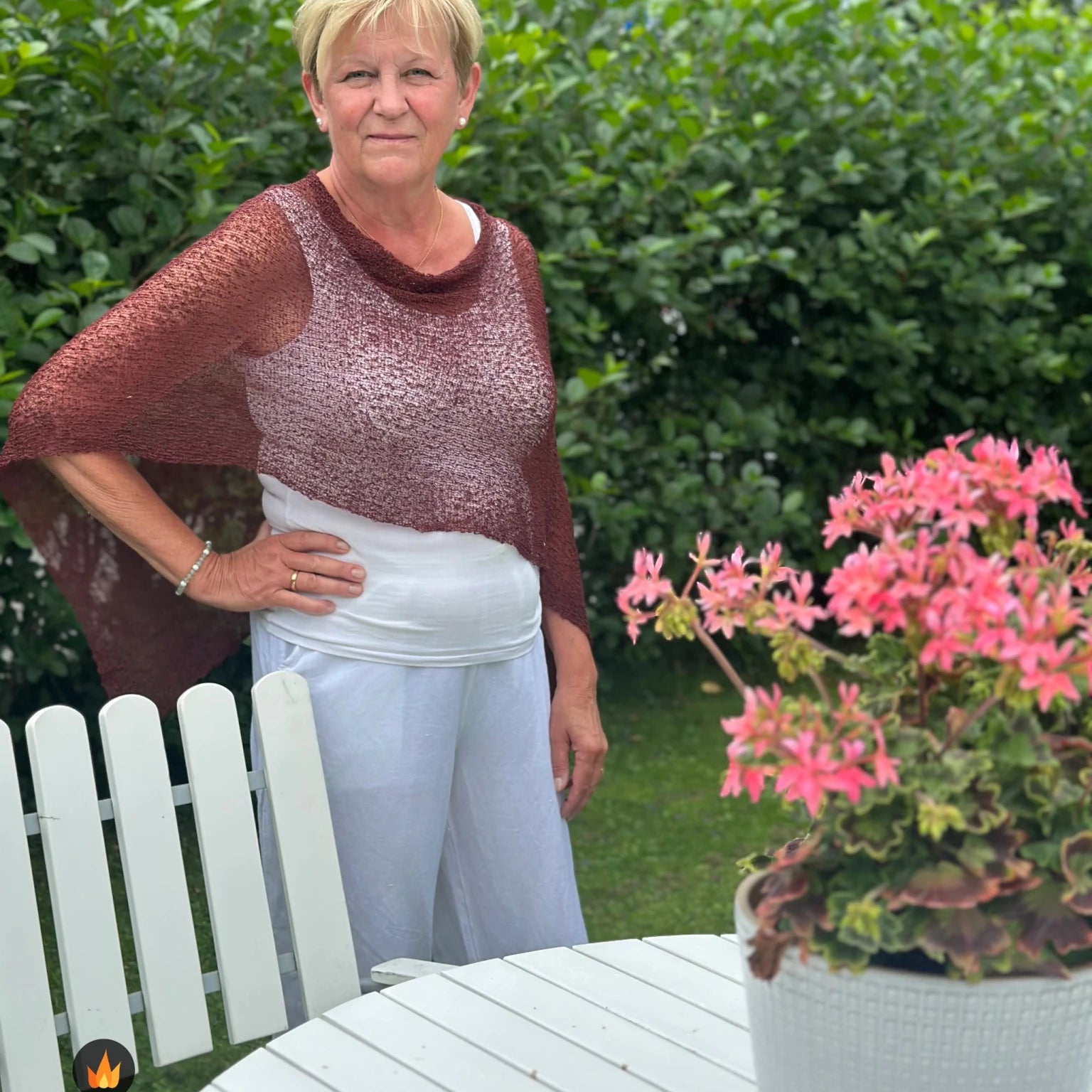 Kvinna bär Ella Cape i färg 026 Rödbrun, en varm färg i kastanjebrun med röd glöd, finstickad cape som draperas mjukt över axlarna, fotad i en sommarträdgård. Ulla Jacobsson Kläder.