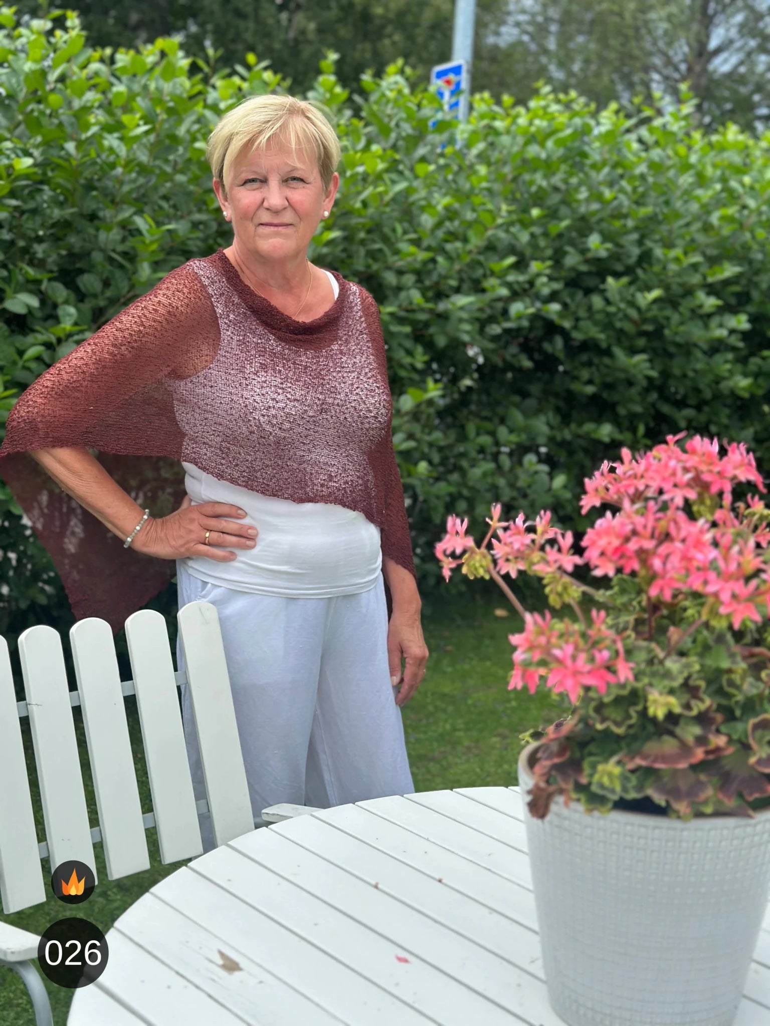 Kvinna bär Ella Cape i färg 026 Rödbrun, en varm färg i kastanjebrun med röd glöd, finstickad cape som draperas mjukt över axlarna, fotad i en sommarträdgård. Ulla Jacobsson Kläder.
