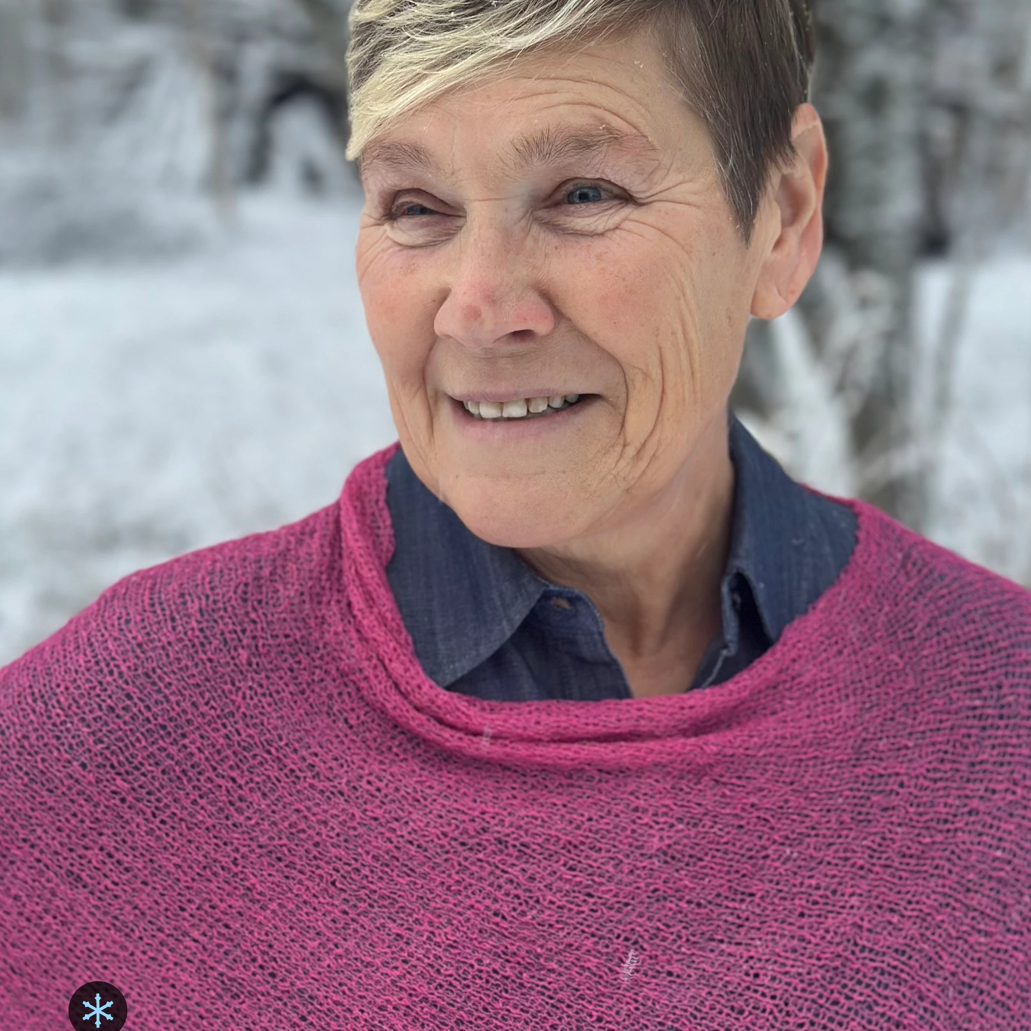 Kvinna bär Ella Cape i färg 027 Rosa, en kall färg i klar rosa, finstickad cape som draperas mjukt över axlarna, fotad i ett snöigt vinterlandskap. Ulla Jacobsson Kläder.