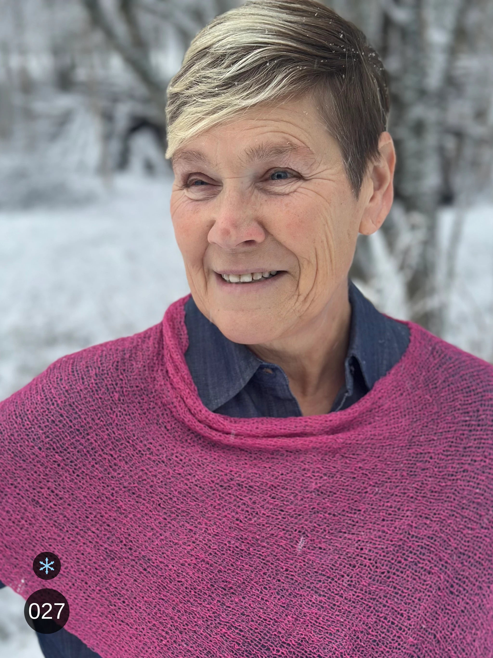 Kvinna bär Ella Cape i färg 027 Rosa, en kall färg i klar rosa, finstickad cape som draperas mjukt över axlarna, fotad i ett snöigt vinterlandskap. Ulla Jacobsson Kläder.