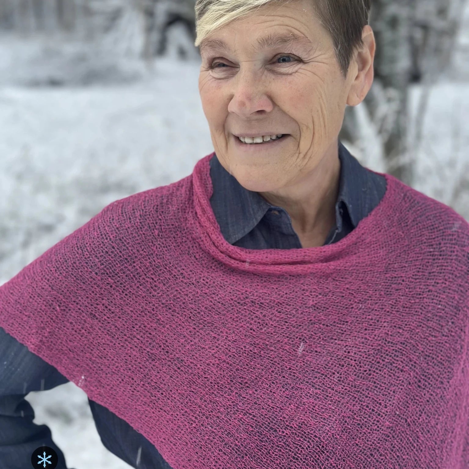Kvinna bär Ella Cape i färg 027 Rosa, en kall färg, finstickad cape som draperas mjukt över axlarna, fotad i ett snöigt vinterlandskap. Ulla Jacobsson Kläder.