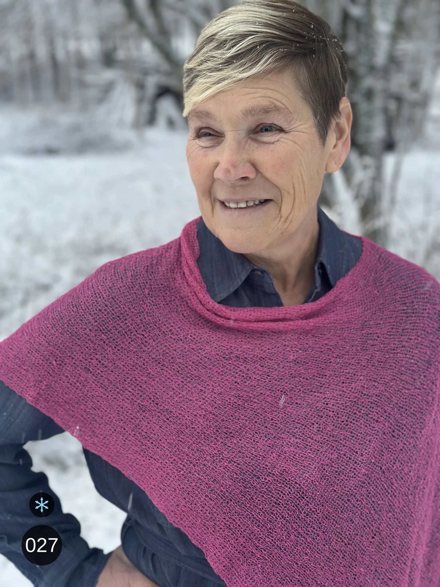 Kvinna bär Ella Cape i färg 027 Rosa, en kall färg, finstickad cape som draperas mjukt över axlarna, fotad i ett snöigt vinterlandskap. Ulla Jacobsson Kläder.