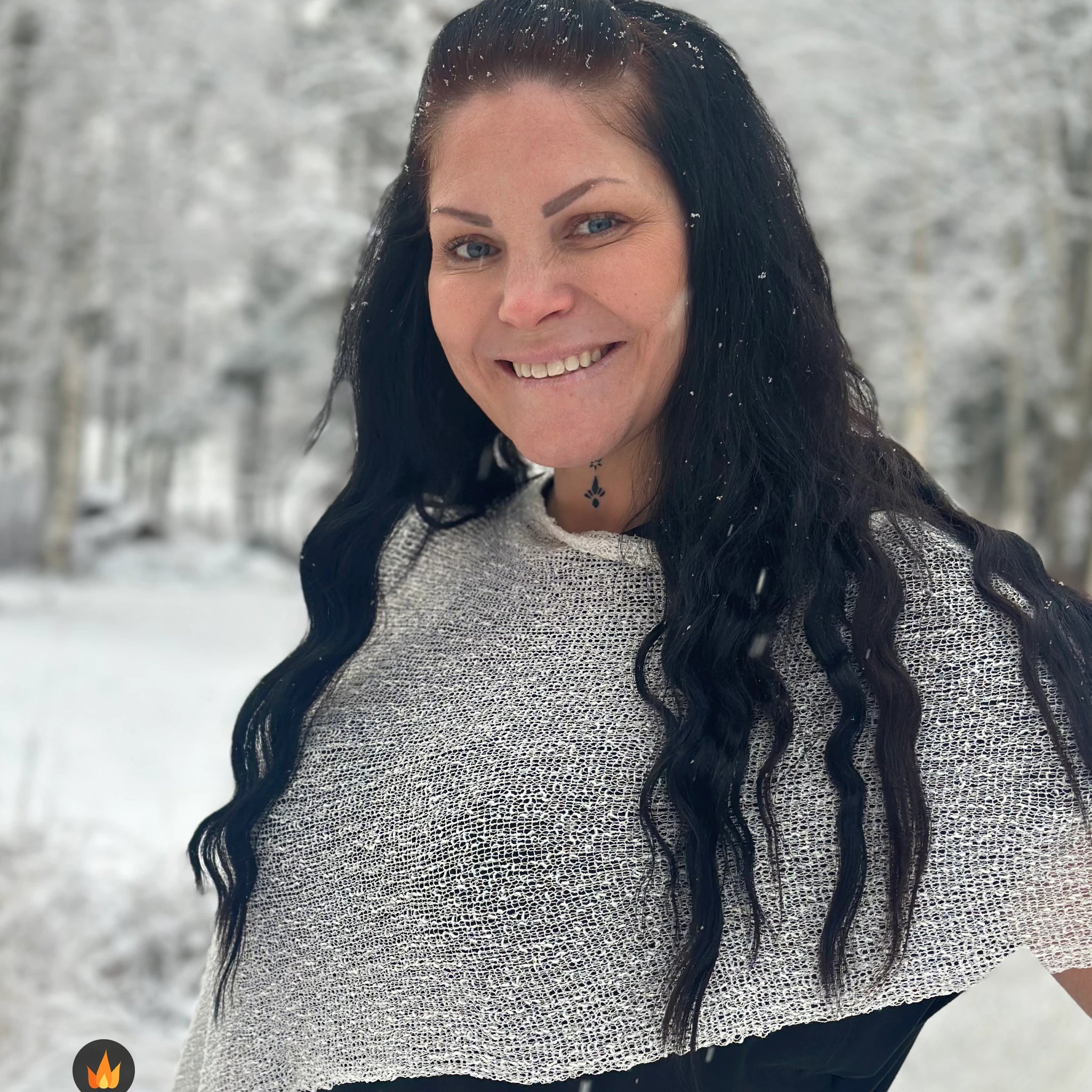 Kvinna bär Ella Cape i färg 031 Offwhite, en varm färg i mjuk offwhite, finstickad cape som draperas mjukt över axlarna, fotad bland snöiga träd. Ulla Jacobsson Kläder.