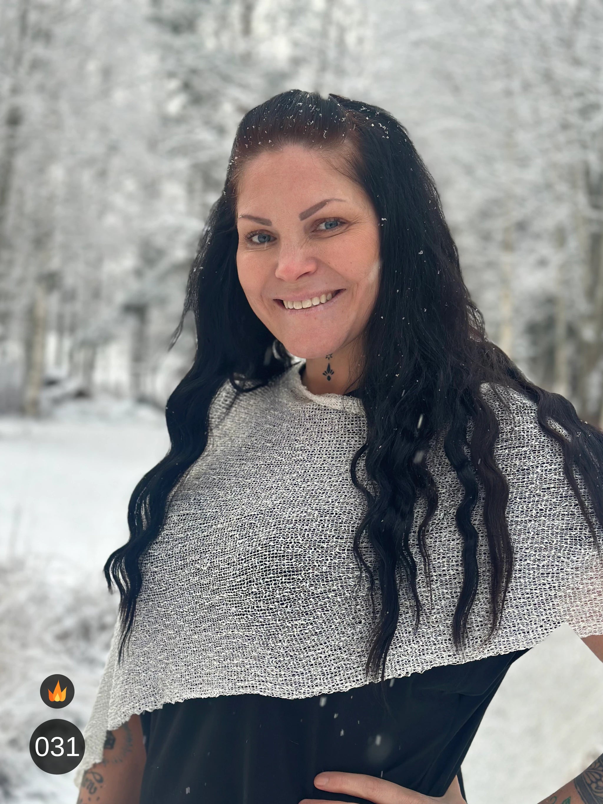 Kvinna bär Ella Cape i färg 031 Offwhite, en varm färg i mjuk offwhite, finstickad cape som draperas mjukt över axlarna, fotad bland snöiga träd. Ulla Jacobsson Kläder.
