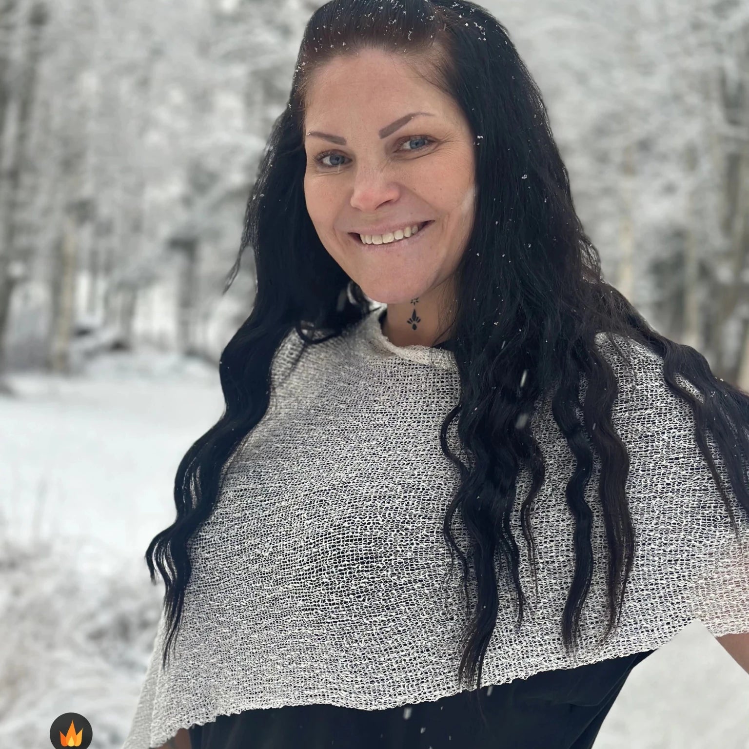 Kvinna bär Ella Cape i färg 031 Offwhite, en varm färg, finstickad cape som draperas mjukt över axlarna, fotad bland snöiga träd. Ulla Jacobsson Kläder.