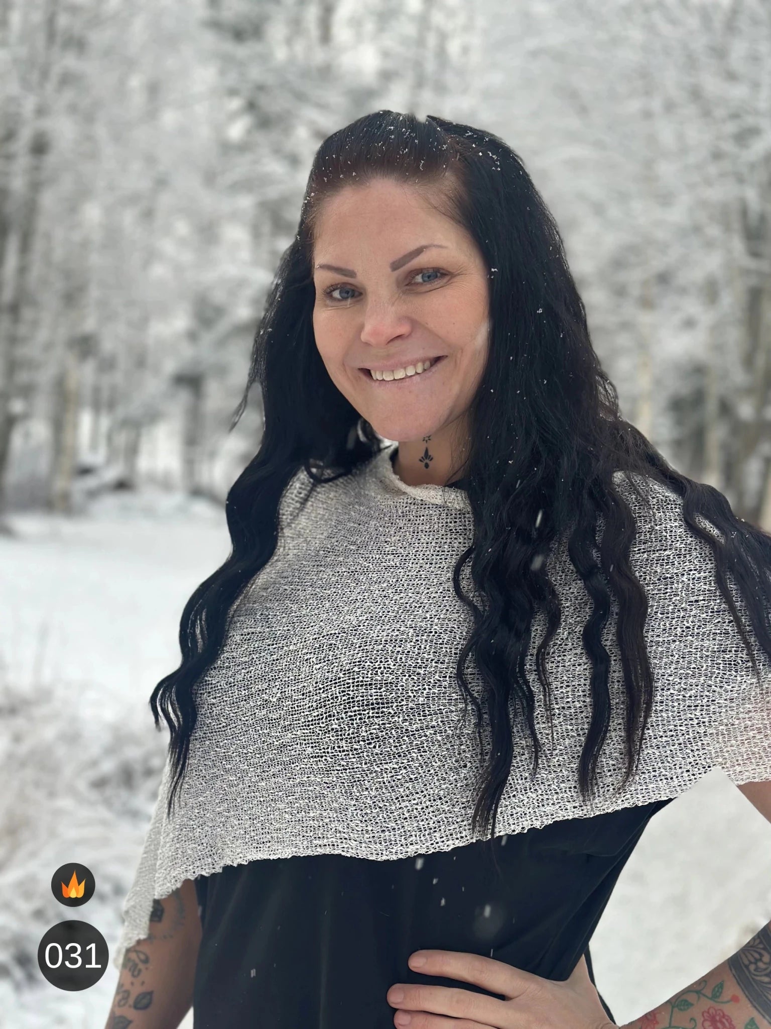 Kvinna bär Ella Cape i färg 031 Offwhite, en varm färg, finstickad cape som draperas mjukt över axlarna, fotad bland snöiga träd. Ulla Jacobsson Kläder.