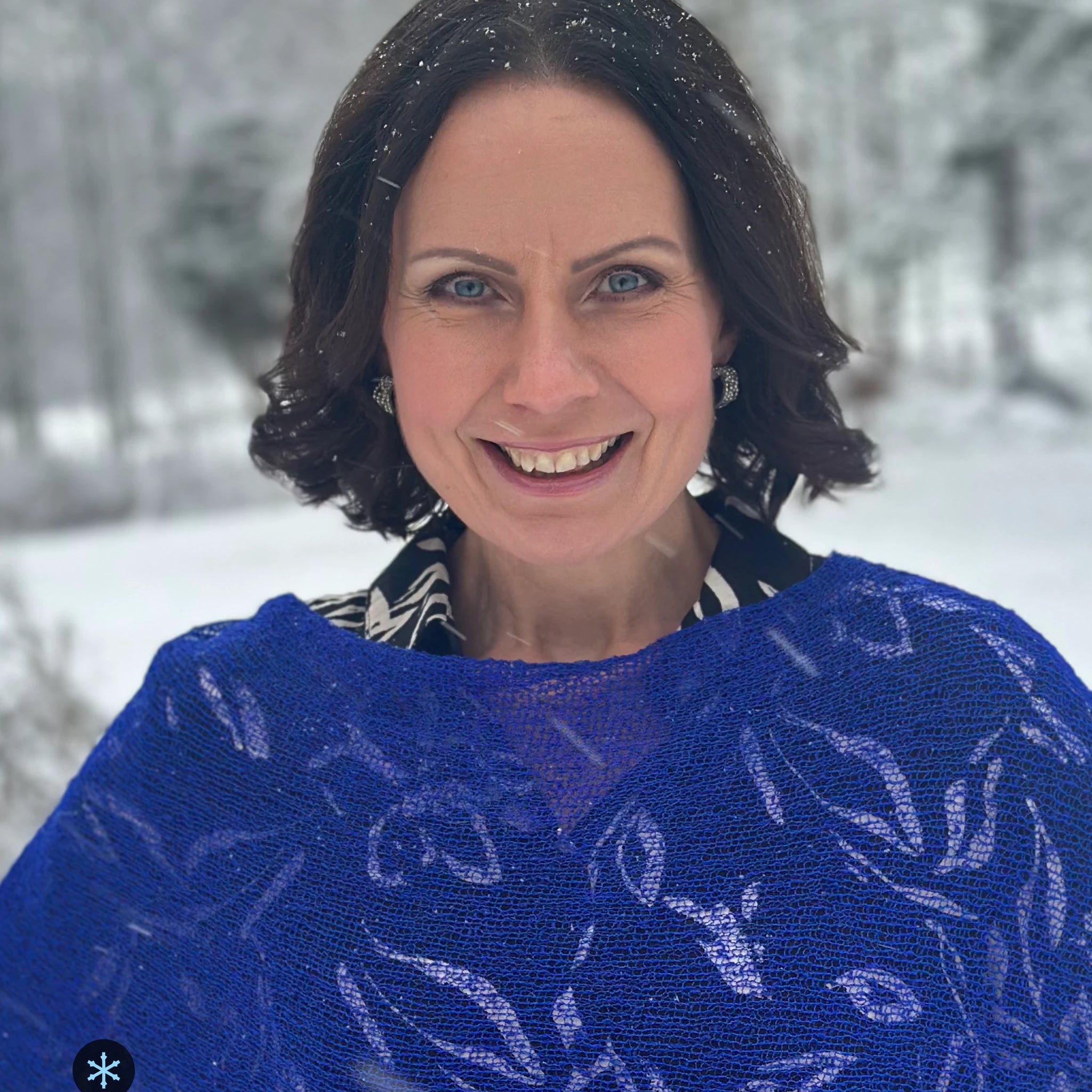 Kvinna bär Ella Cape i färg 032 Klarblå, en kall färg i intensiv klarblå, finstickad cape som draperas mjukt över axlarna, fotad en snöig vinterdag. Ulla Jacobsson Kläder.
