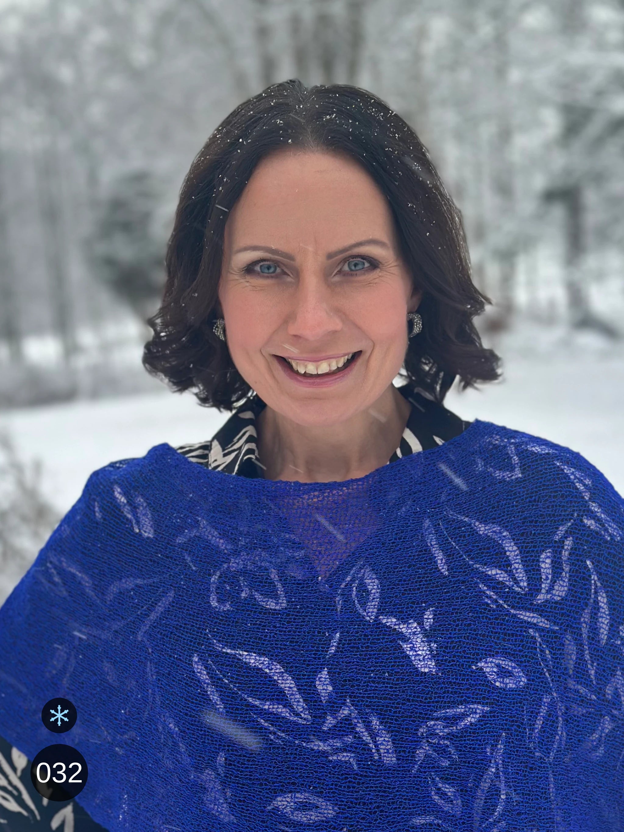 Kvinna bär Ella Cape i färg 032 Klarblå, en kall färg i intensiv klarblå, finstickad cape som draperas mjukt över axlarna, fotad en snöig vinterdag. Ulla Jacobsson Kläder.