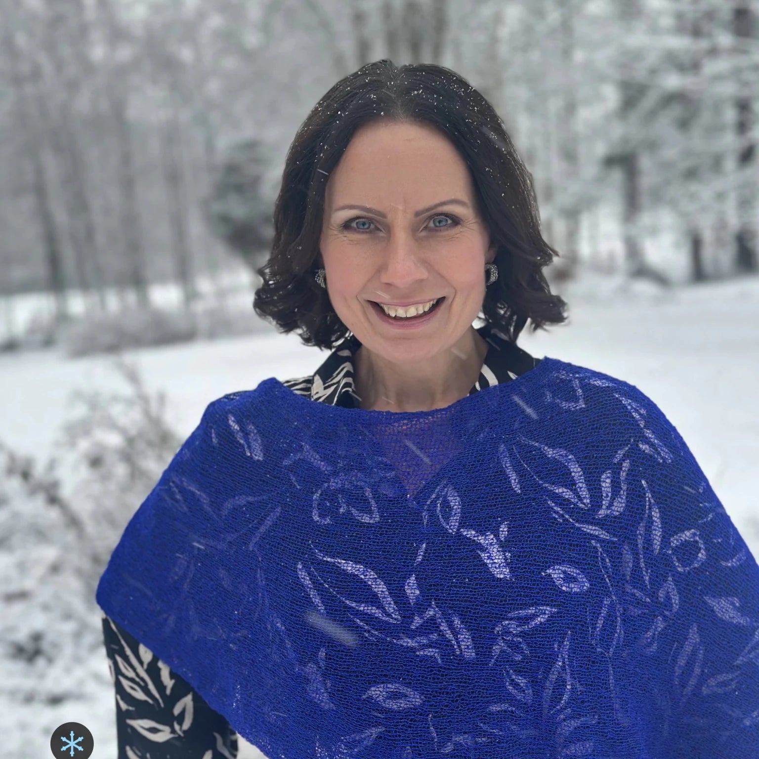 Kvinna bär Ella Cape i färg 032 Klarblå, en kall färg, finstickad cape som draperas mjukt över axlarna, fotad i ett snöigt vinterlandskap. Ulla Jacobsson Kläder.