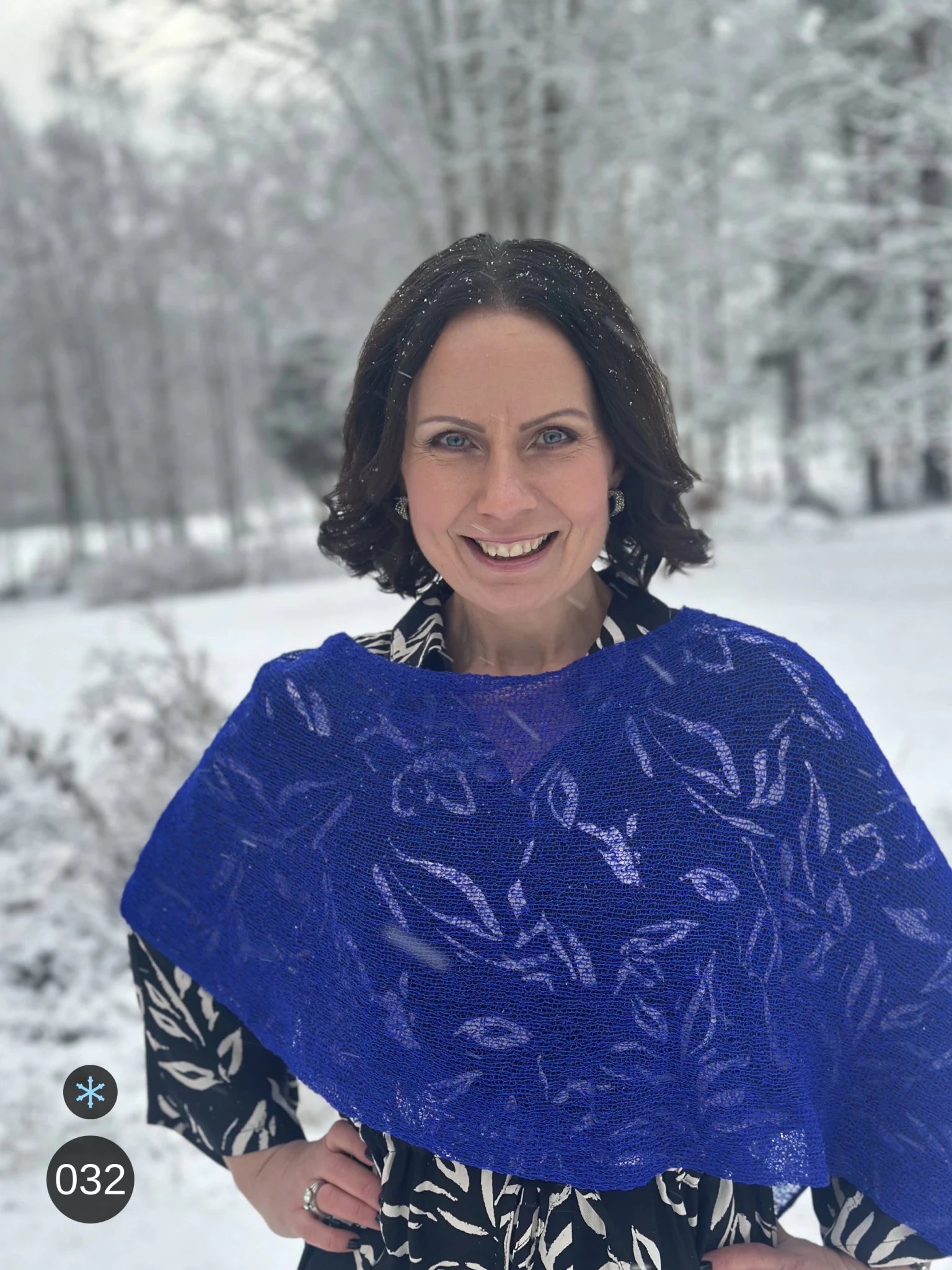 Kvinna bär Ella Cape i färg 032 Klarblå, en kall färg, finstickad cape som draperas mjukt över axlarna, fotad i ett snöigt vinterlandskap. Ulla Jacobsson Kläder.