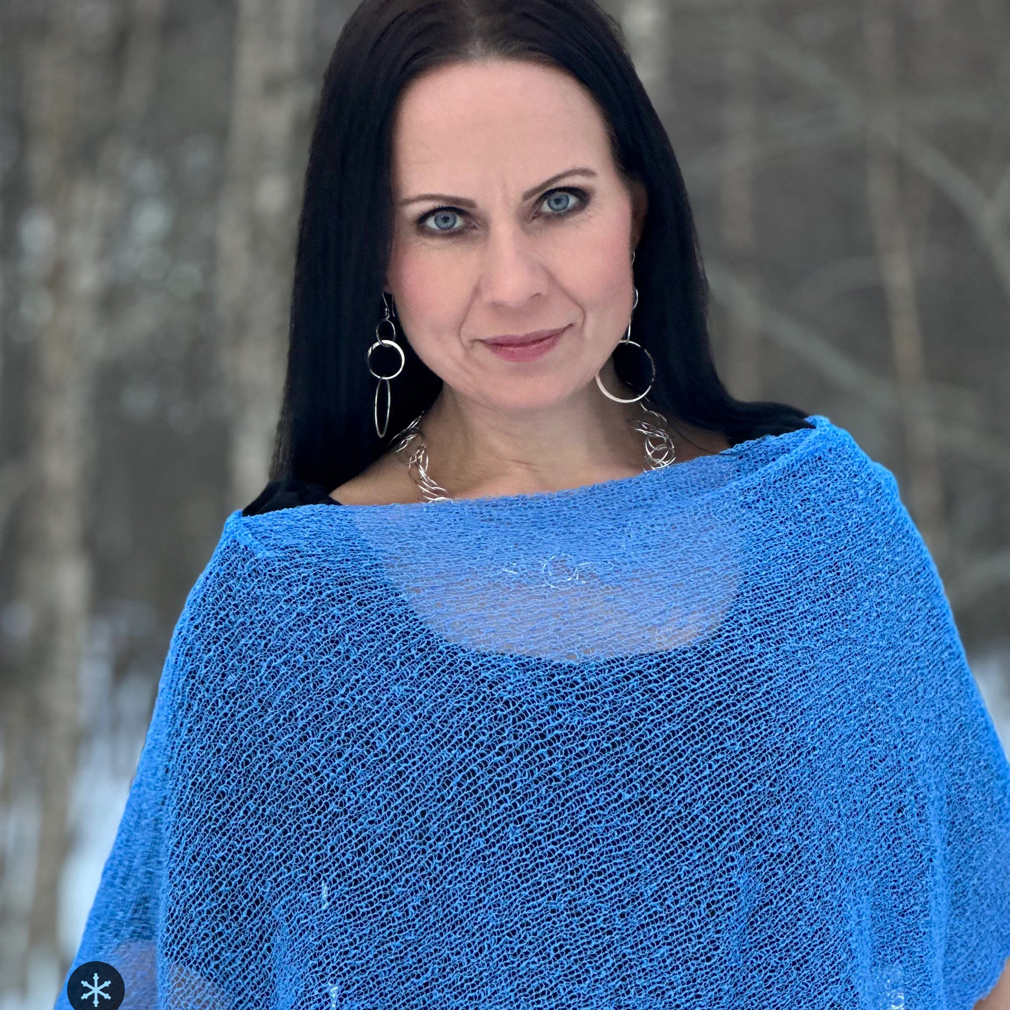Kvinna bär Ella Cape i färg 033 Ljusblå, en kall färg i klar himmelsblå med en aning violett djup, finstickad cape som draperas mjukt över axlarna, fotad bland snöiga björkar. Ulla Jacobsson Kläder.