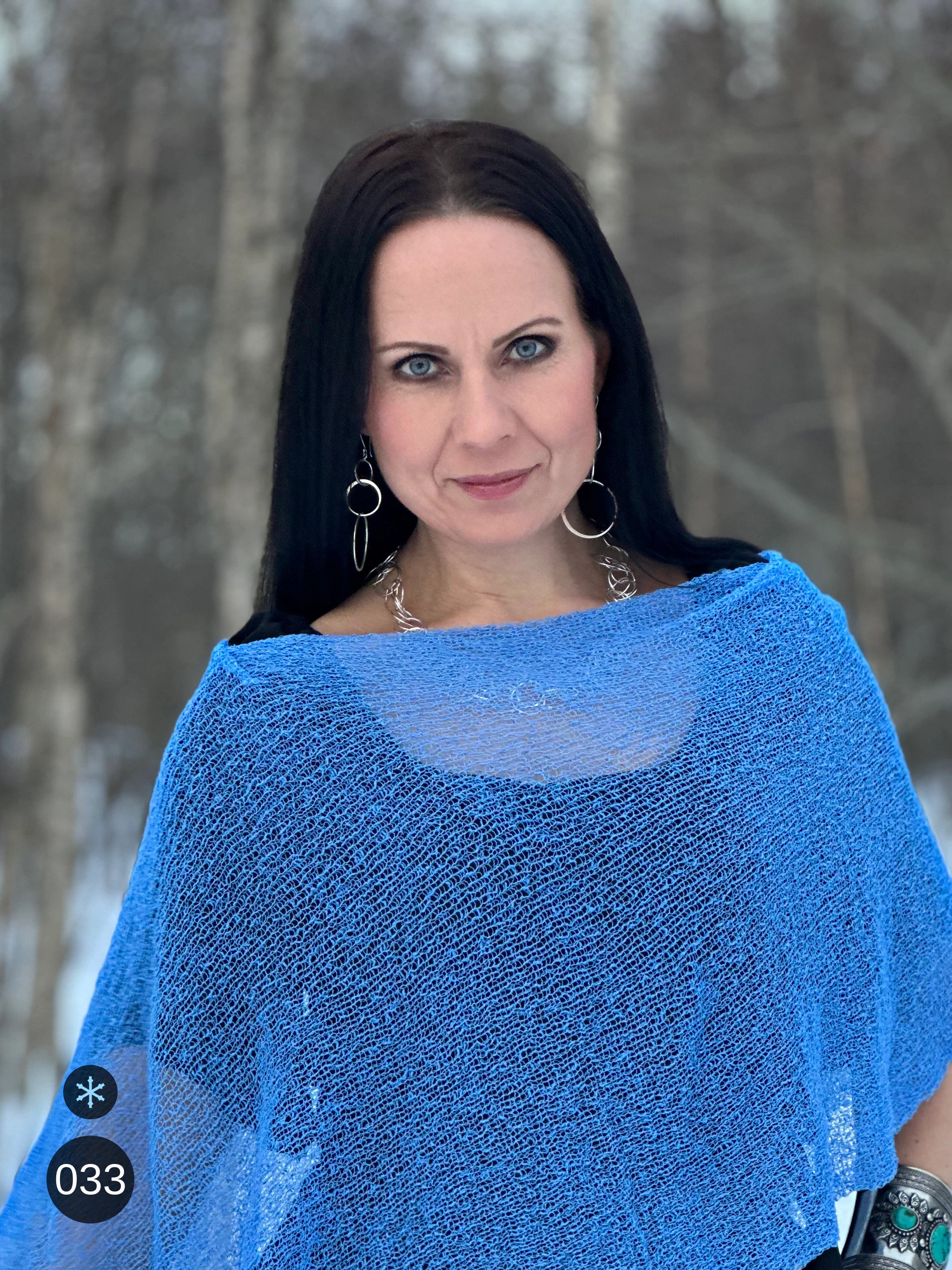 Kvinna bär Ella Cape i färg 033 Ljusblå, en kall färg i klar himmelsblå med en aning violett djup, finstickad cape som draperas mjukt över axlarna, fotad bland snöiga björkar. Ulla Jacobsson Kläder.