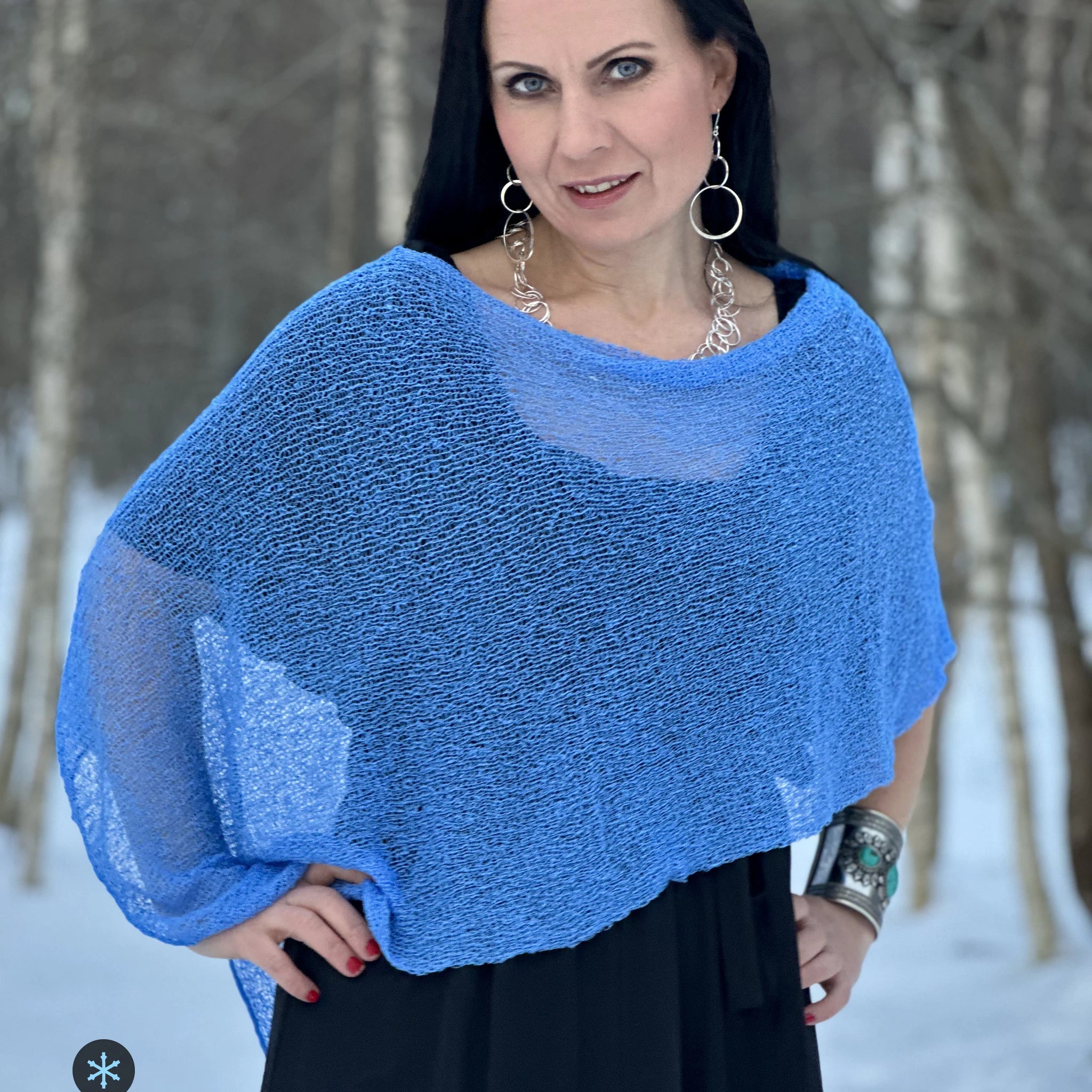 Kvinna bär Ella Cape i färg 033 Ljusblå, en kall färg i klar himmelsblå med en aning violett djup, finstickad cape som draperas mjukt över axlarna, fotad bland snöiga björkar. Ulla Jacobsson Kläder.