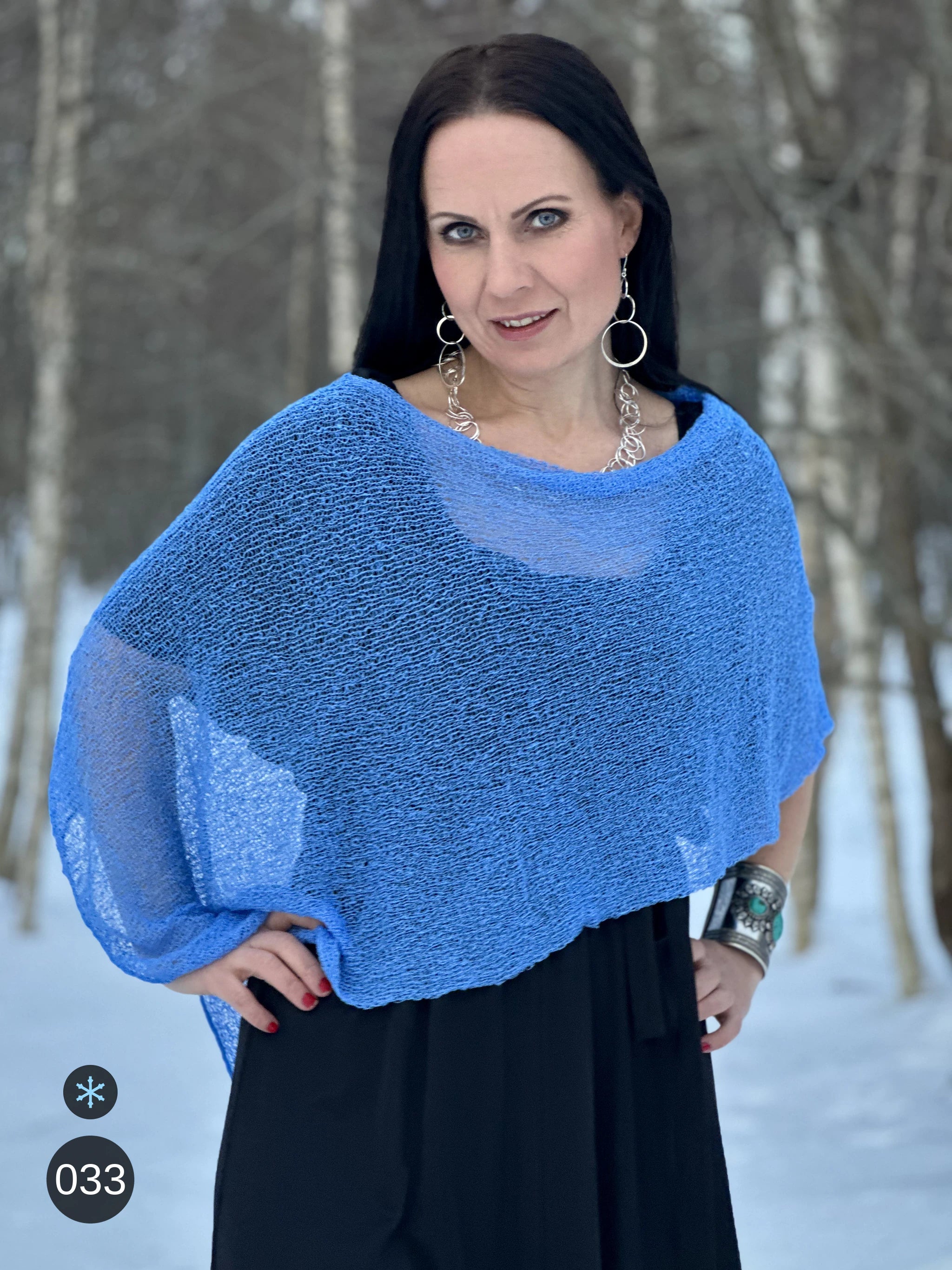 Kvinna bär Ella Cape i färg 033 Ljusblå, en kall färg i klar himmelsblå med en aning violett djup, finstickad cape som draperas mjukt över axlarna, fotad bland snöiga björkar. Ulla Jacobsson Kläder.