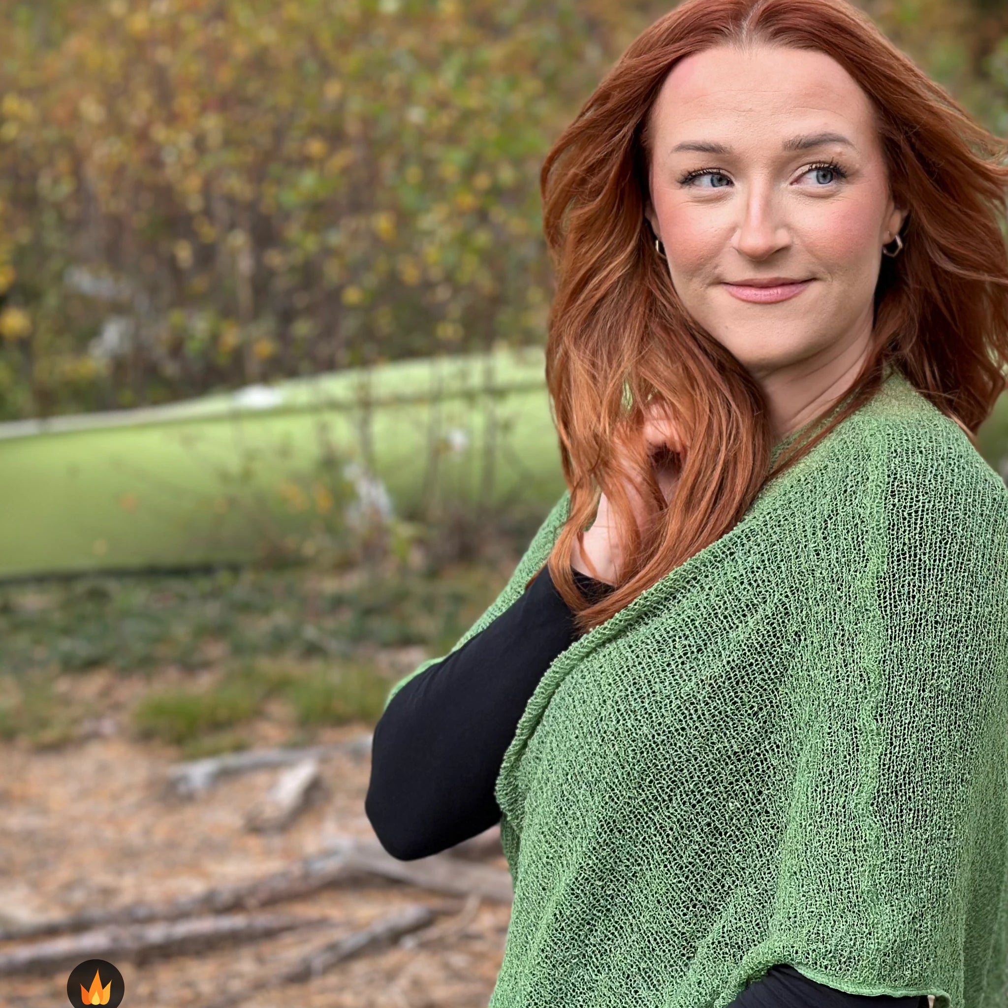 Kvinna bär Ella Cape i färg 034 Ljusgrön, en varm färg i ljus vårgrön, finstickad cape som draperas mjukt över axlarna, fotad utomhus i grönska. Ulla Jacobsson Kläder.