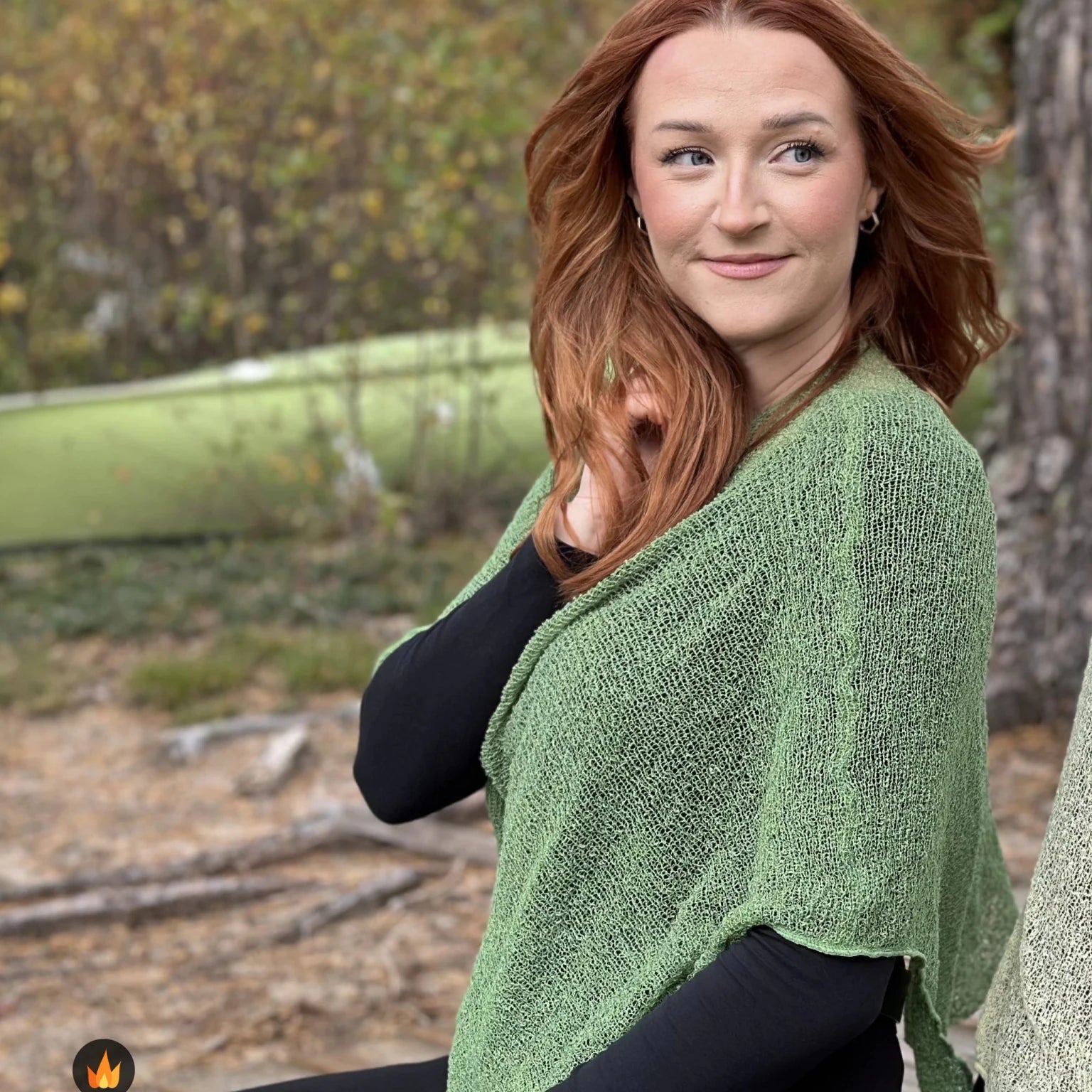 Kvinna bär Ella Cape i färg 034 Ljusgrön, en varm färg, finstickad cape som draperas mjukt över axlarna, fotad utomhus i grönska. Ulla Jacobsson Kläder.