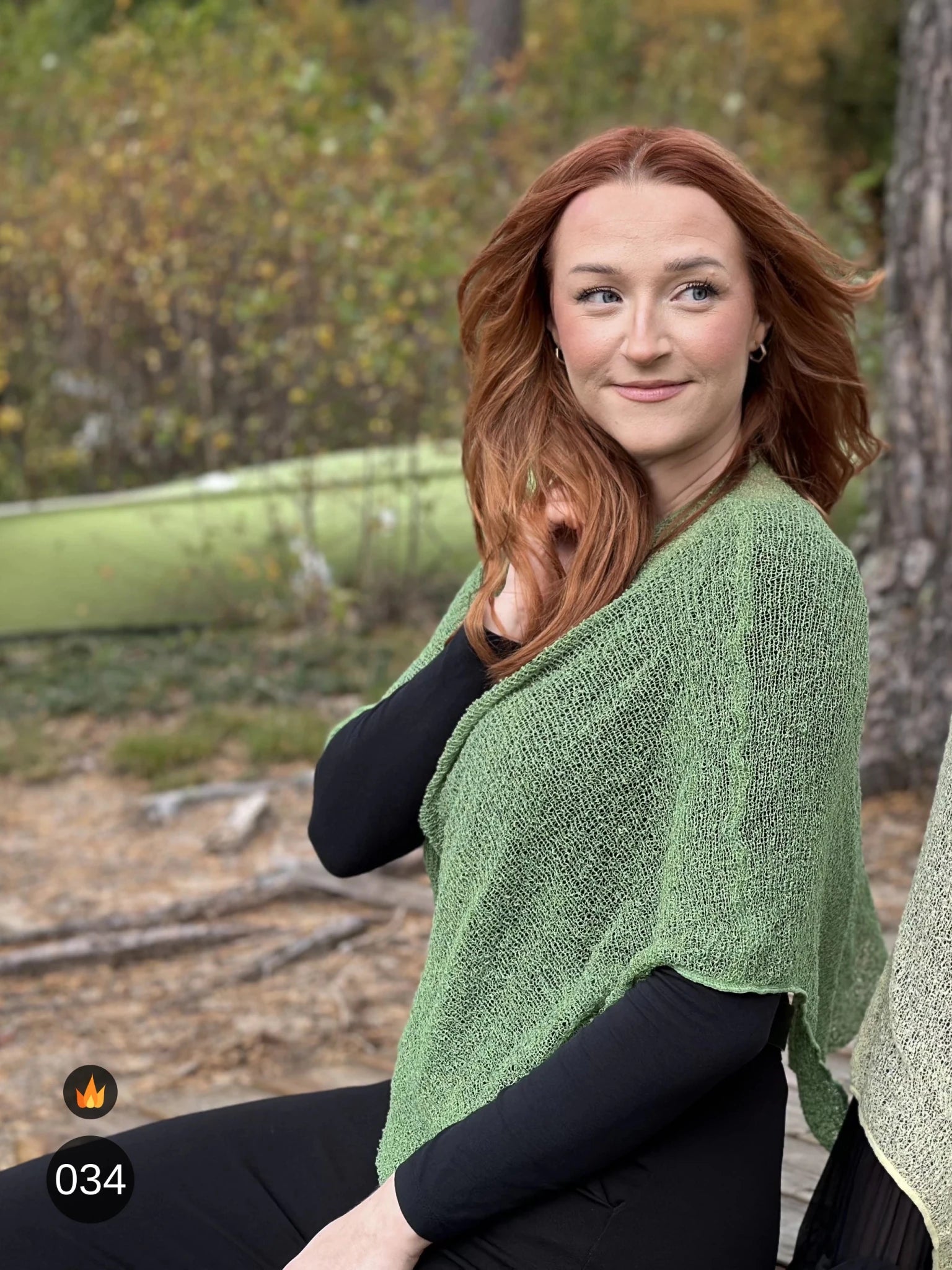 Kvinna bär Ella Cape i färg 034 Ljusgrön, en varm färg, finstickad cape som draperas mjukt över axlarna, fotad utomhus i grönska. Ulla Jacobsson Kläder.