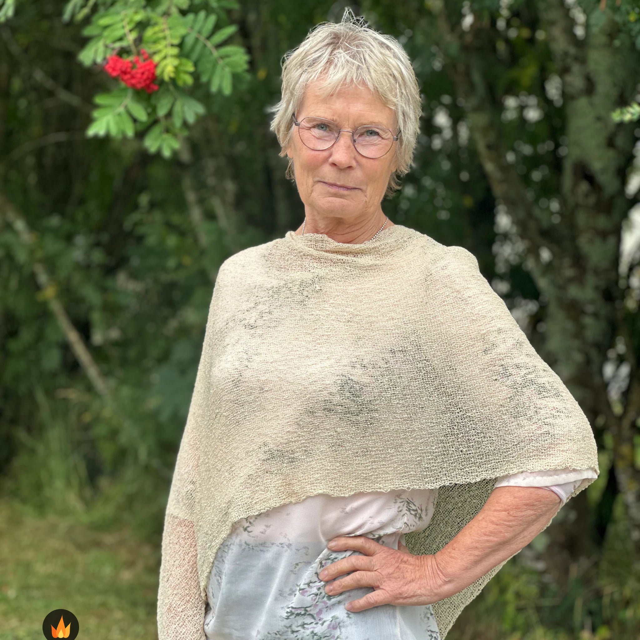 Kvinna bär Ella Cape i färg 036 Ljus sand, en varm färg i ljus sandbeige med champagneton, finstickad cape som draperas mjukt över axlarna, fotad utomhus i grönska. Ulla Jacobsson Kläder.