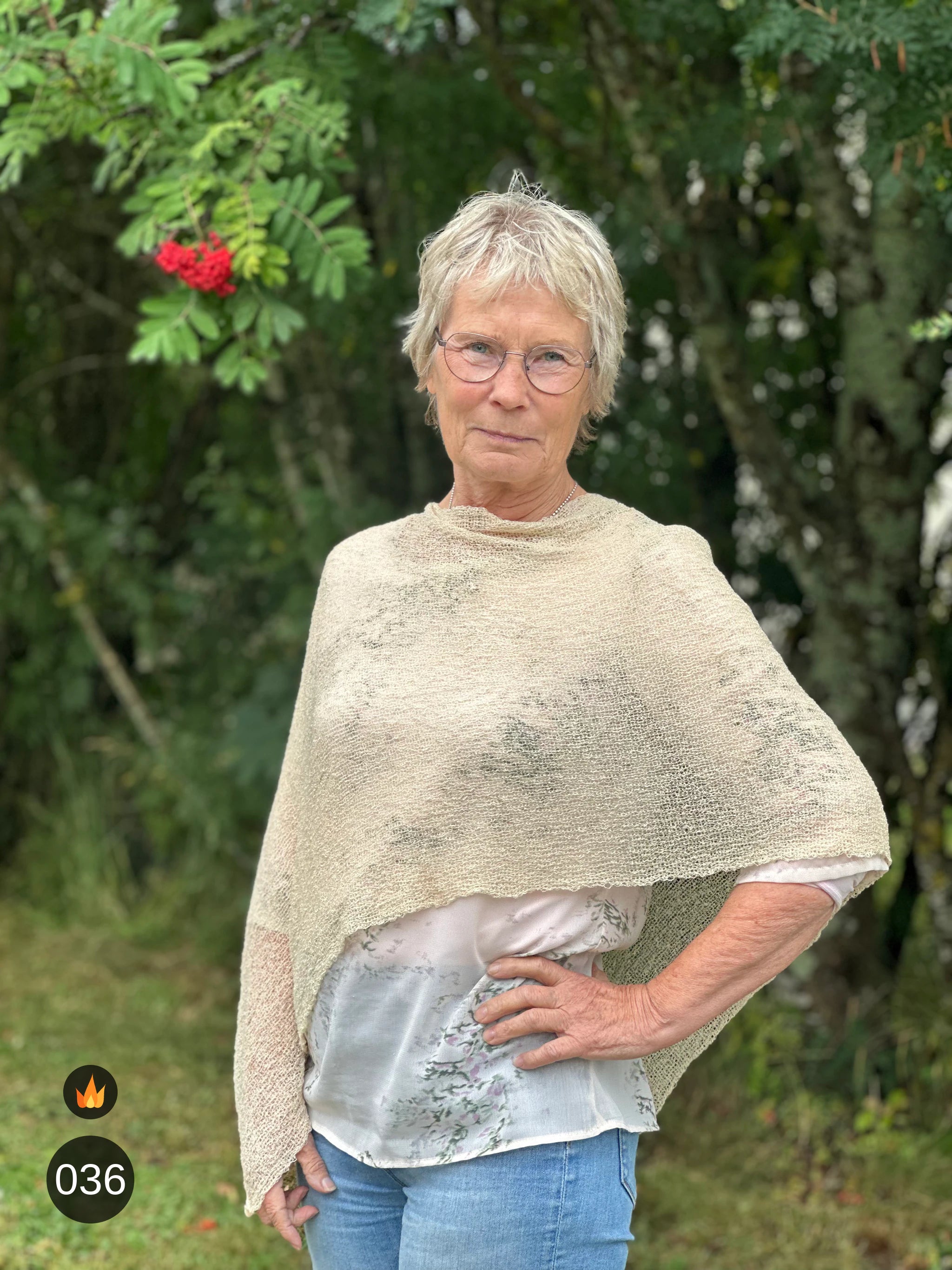 Kvinna bär Ella Cape i färg 036 Ljus sand, en varm färg i ljus sandbeige med champagneton, finstickad cape som draperas mjukt över axlarna, fotad utomhus i grönska. Ulla Jacobsson Kläder.