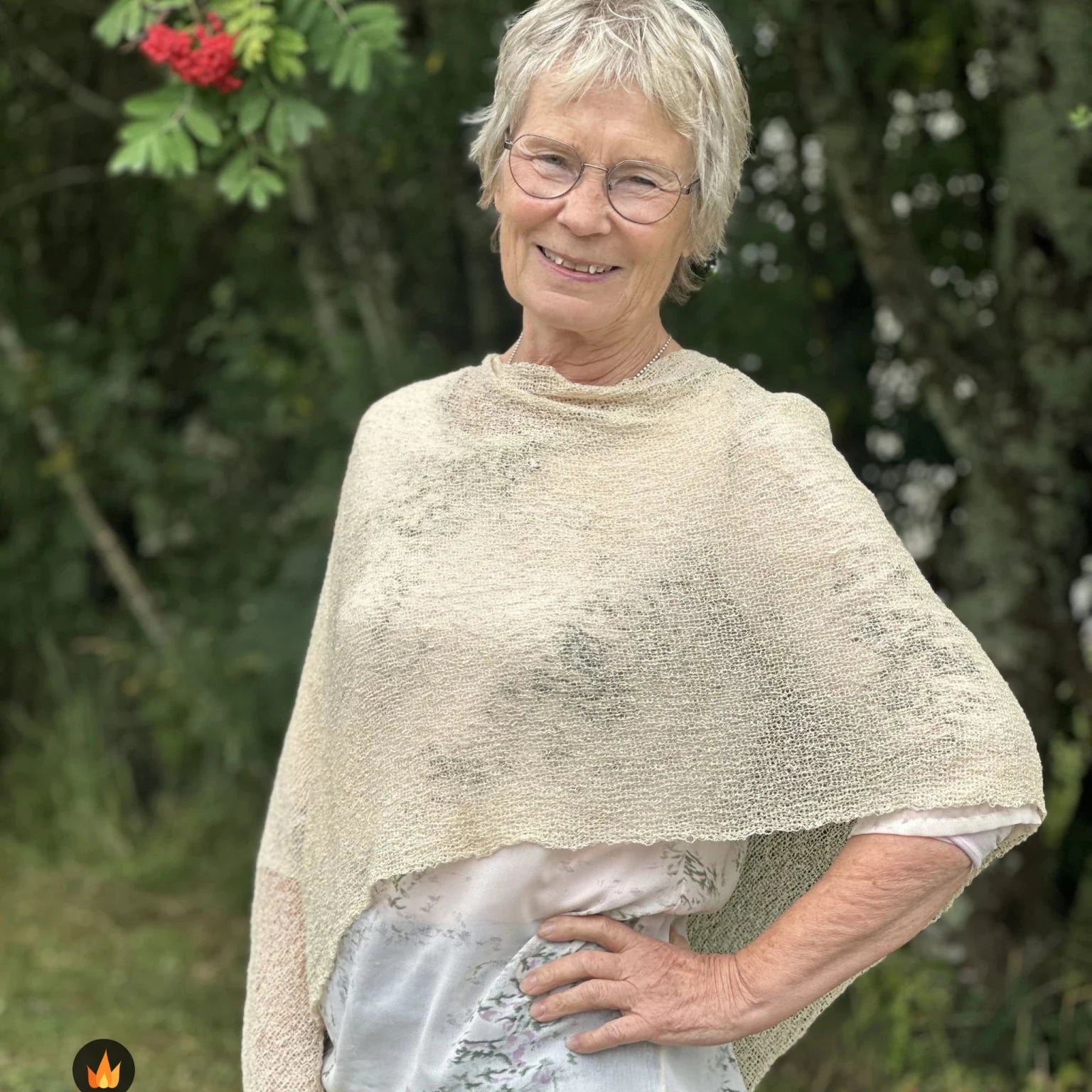 Kvinna bär Ella Cape i färg 036 Ljus sand, en varm färg, finstickad cape som draperas mjukt över axlarna, fotad utomhus i grönska. Ulla Jacobsson Kläder.