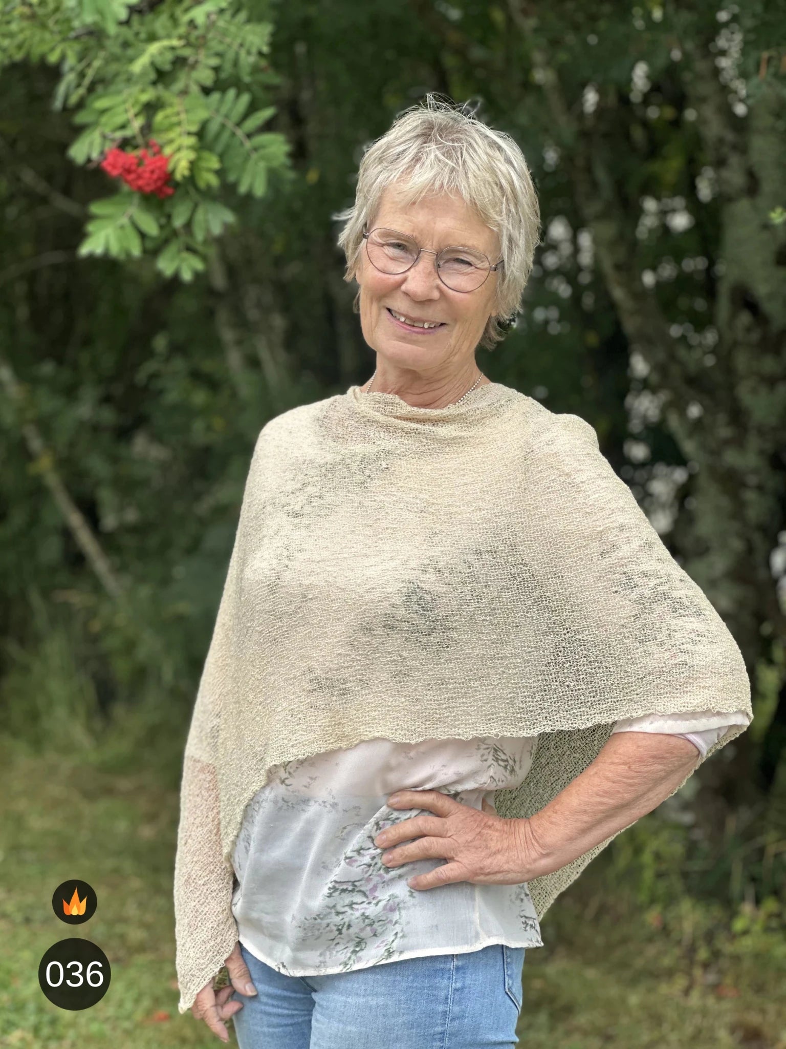 Kvinna bär Ella Cape i färg 036 Ljus sand, en varm färg, finstickad cape som draperas mjukt över axlarna, fotad utomhus i grönska. Ulla Jacobsson Kläder.