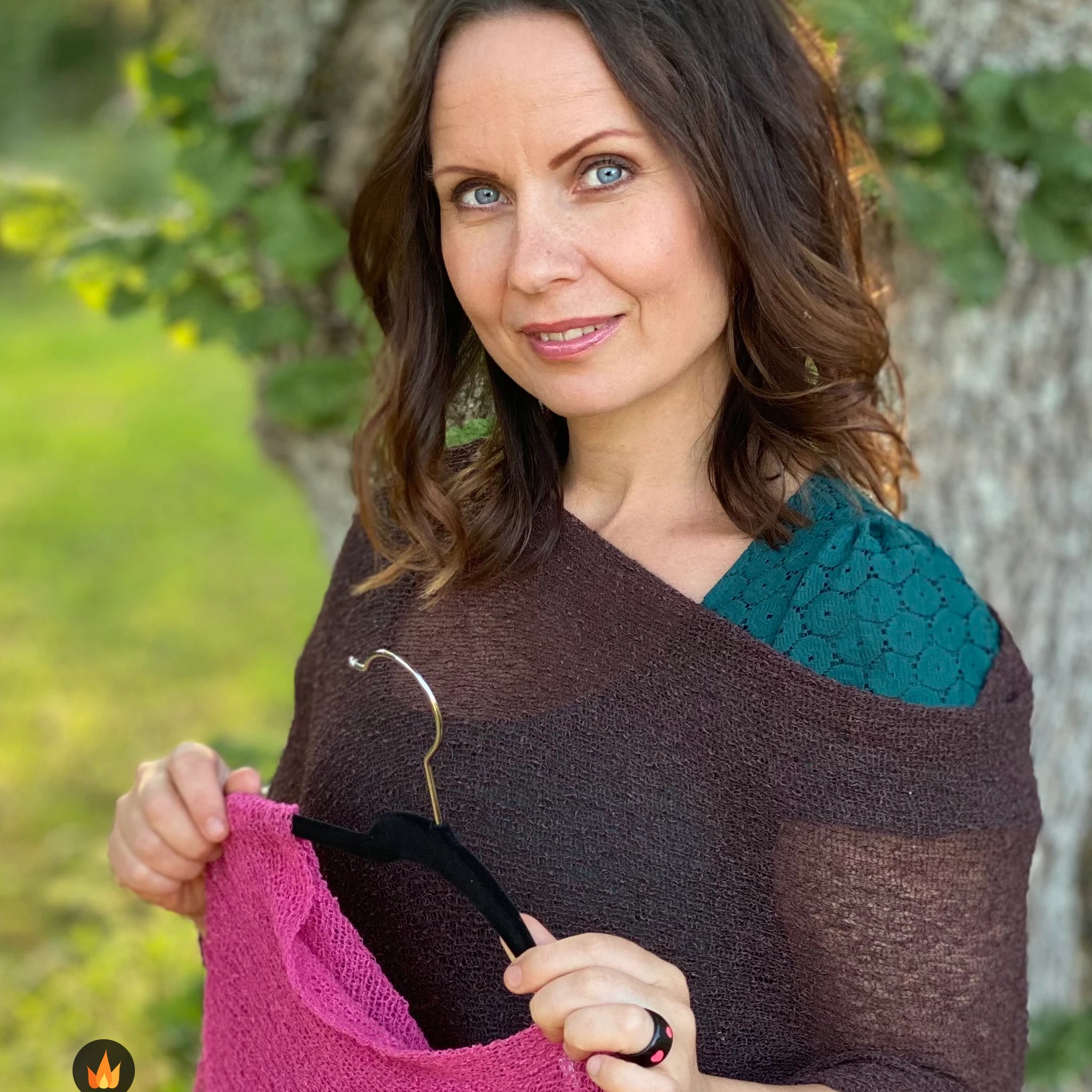 Kvinna bär Ella Cape i färg 037 Mörkbrun, en varm färg i djup mörkbrun, finstickad cape som draperas mjukt över axlarna, fotad i sommargrönska. Ulla Jacobsson Kläder.