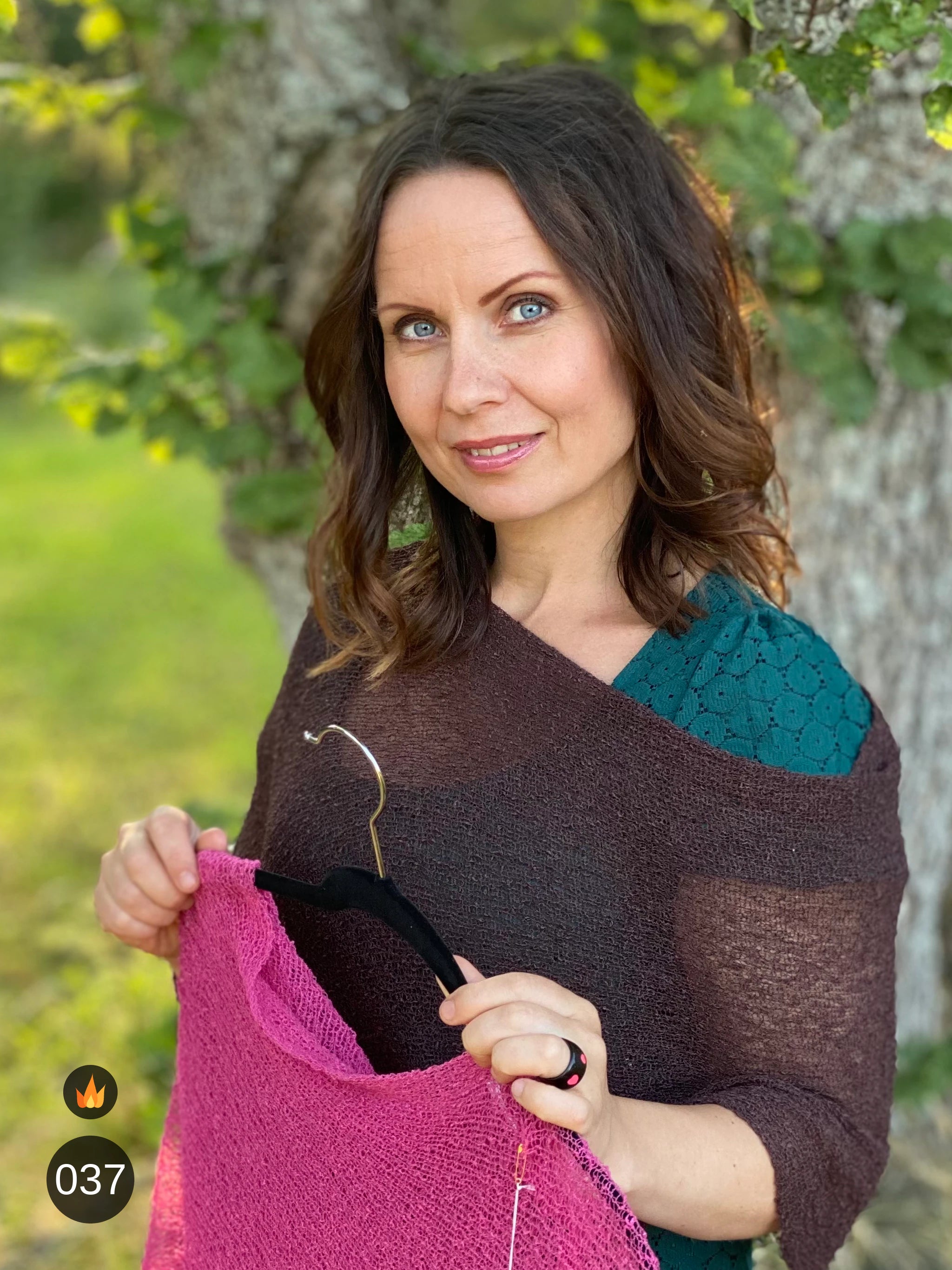 Kvinna bär Ella Cape i färg 037 Mörkbrun, en varm färg i djup mörkbrun, finstickad cape som draperas mjukt över axlarna, fotad i sommargrönska. Ulla Jacobsson Kläder.