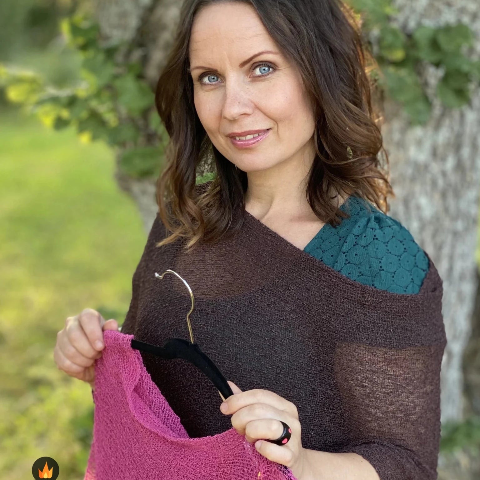 Kvinna bär Ella Cape i färg 037 Mörkbrun, en varm färg, finstickad cape som draperas mjukt över axlarna, fotad utomhus vid blommande träd. Ulla Jacobsson Kläder.