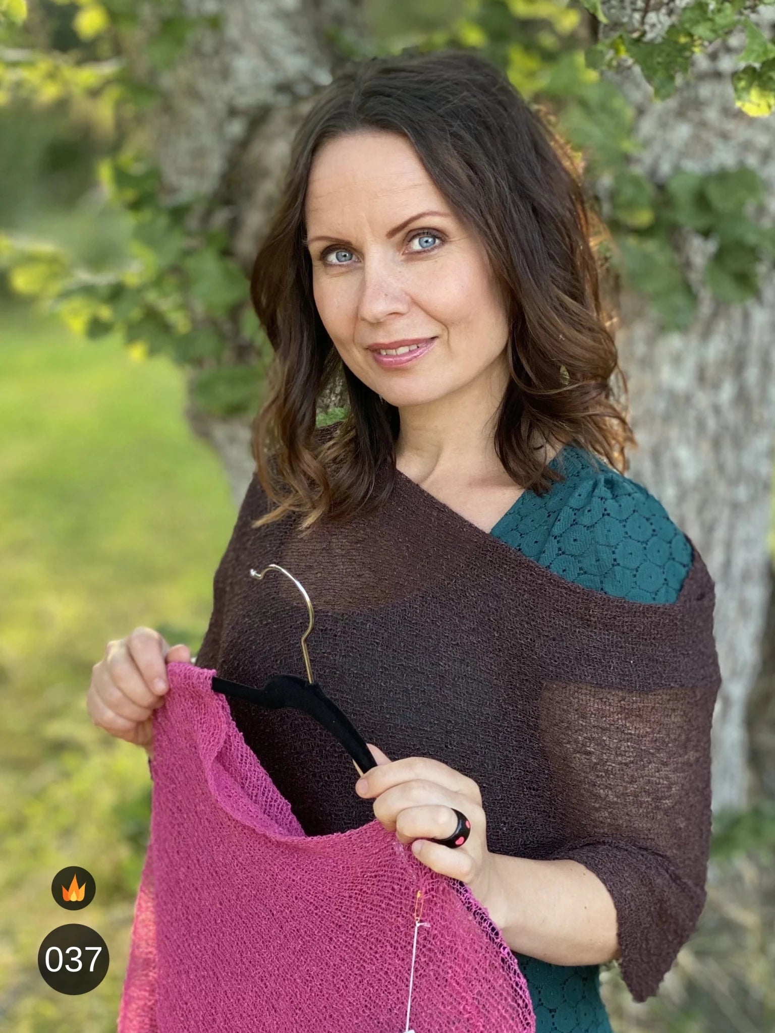 Kvinna bär Ella Cape i färg 037 Mörkbrun, en varm färg, finstickad cape som draperas mjukt över axlarna, fotad utomhus vid blommande träd. Ulla Jacobsson Kläder.