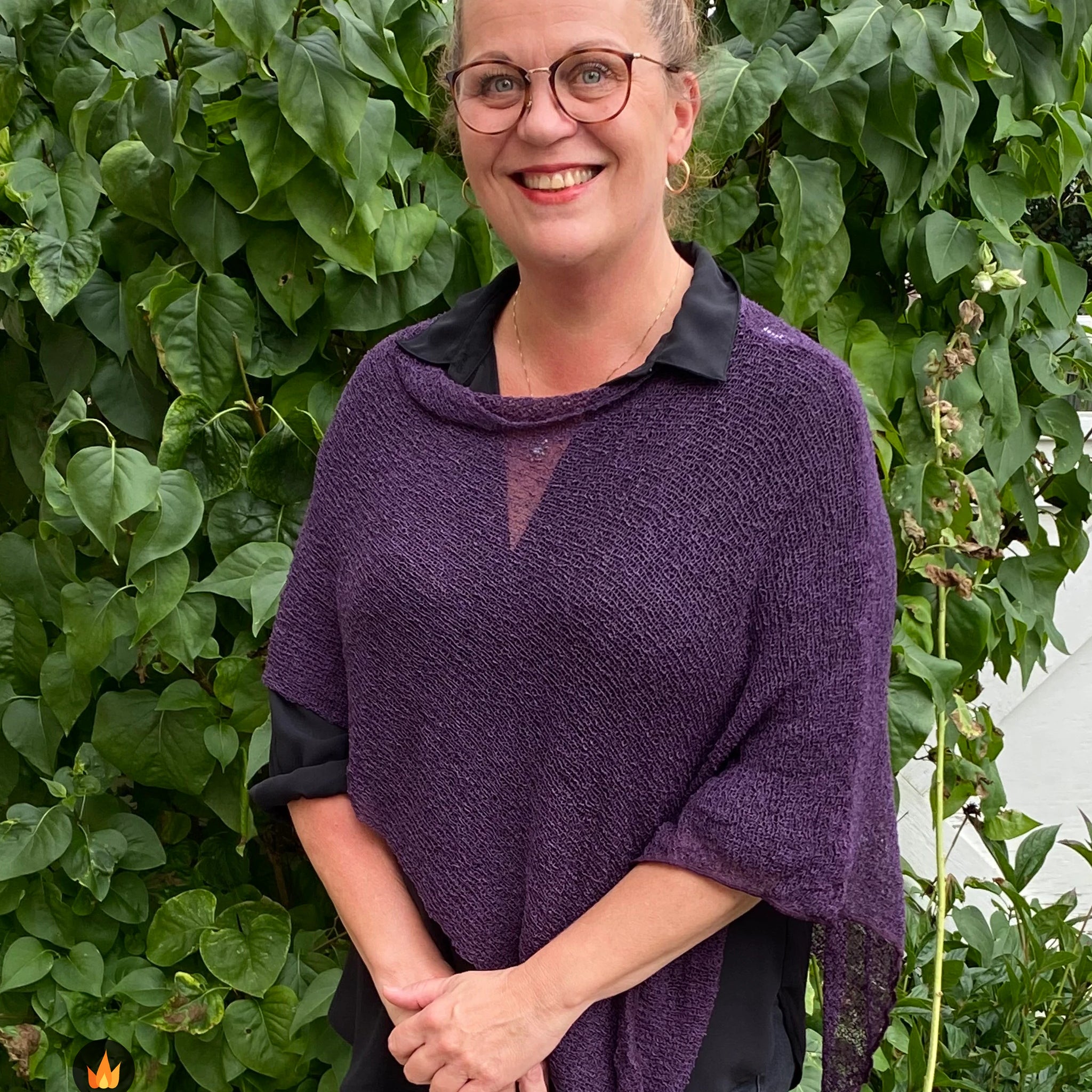 Kvinna bär Ella Cape i färg 038 Aubergine, en varm färg i djup aubergine, finstickad cape som draperas mjukt över axlarna, fotad framför en grön murgrönshäck. Ulla Jacobsson Kläder.